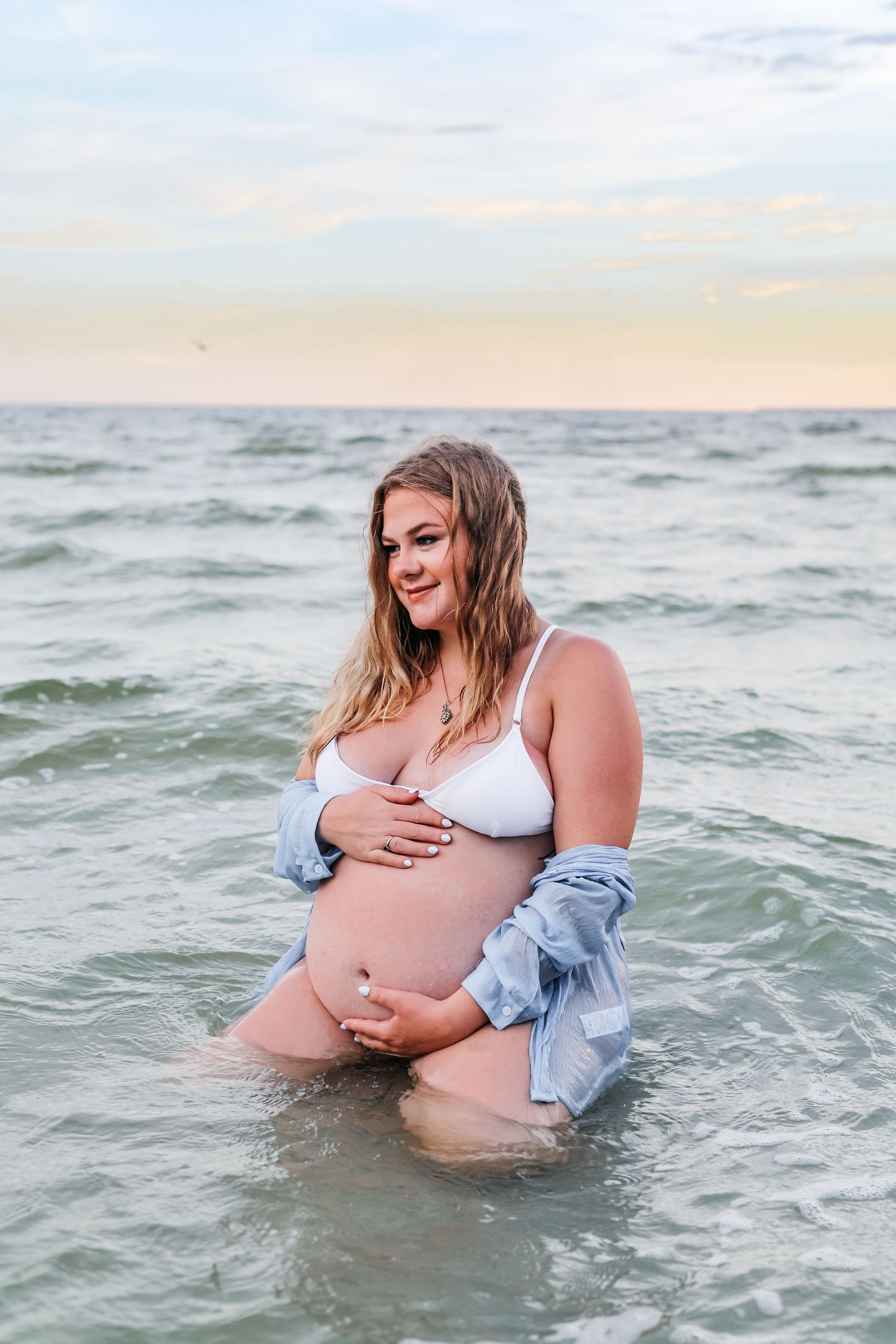 A woman wearing a white bikini and denim jacket poses in ocean waves at sunset during a maternity photoshoot.