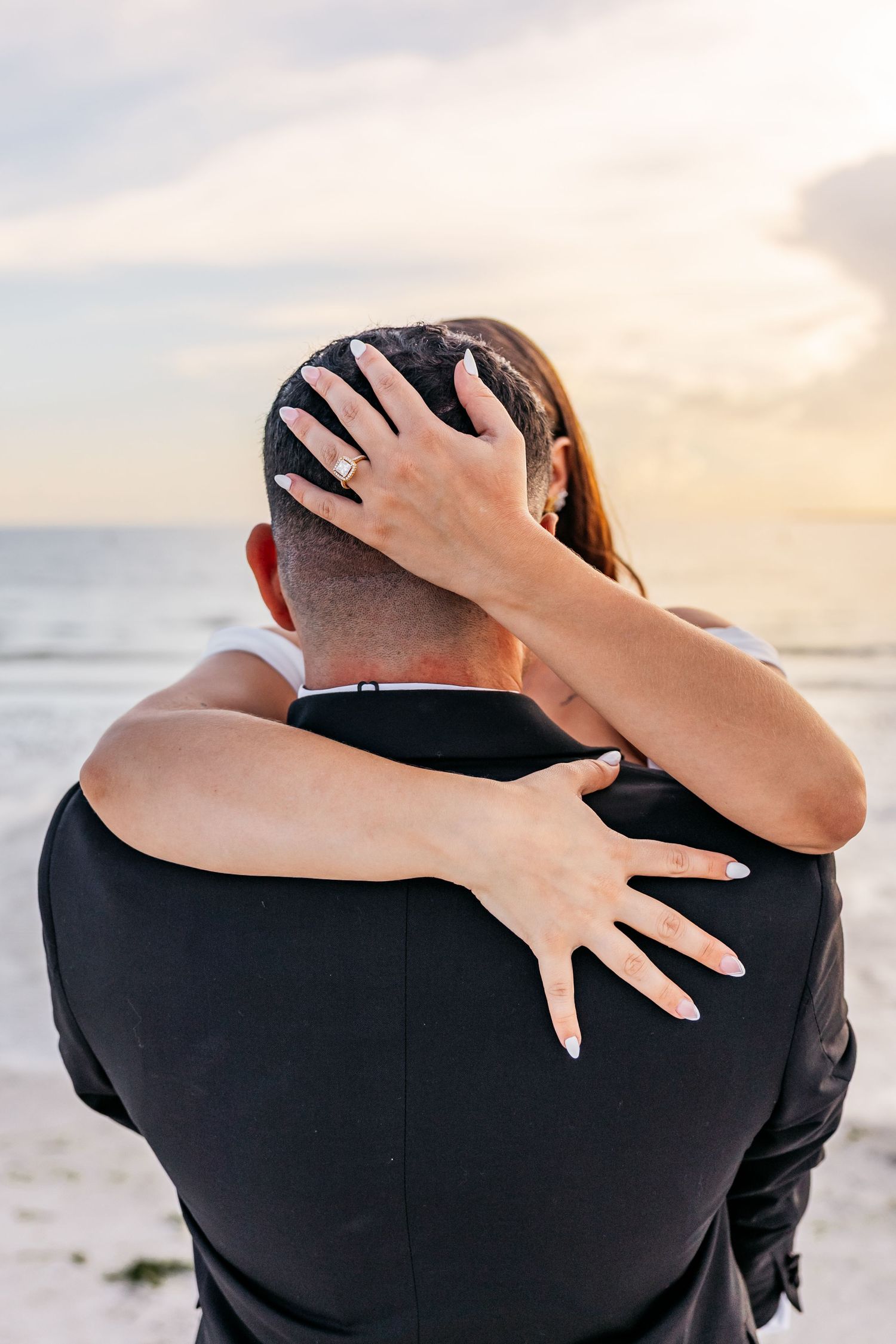 Bride embracing groom from behind on beach at sunset, showcasing elegant diamond engagement ring.