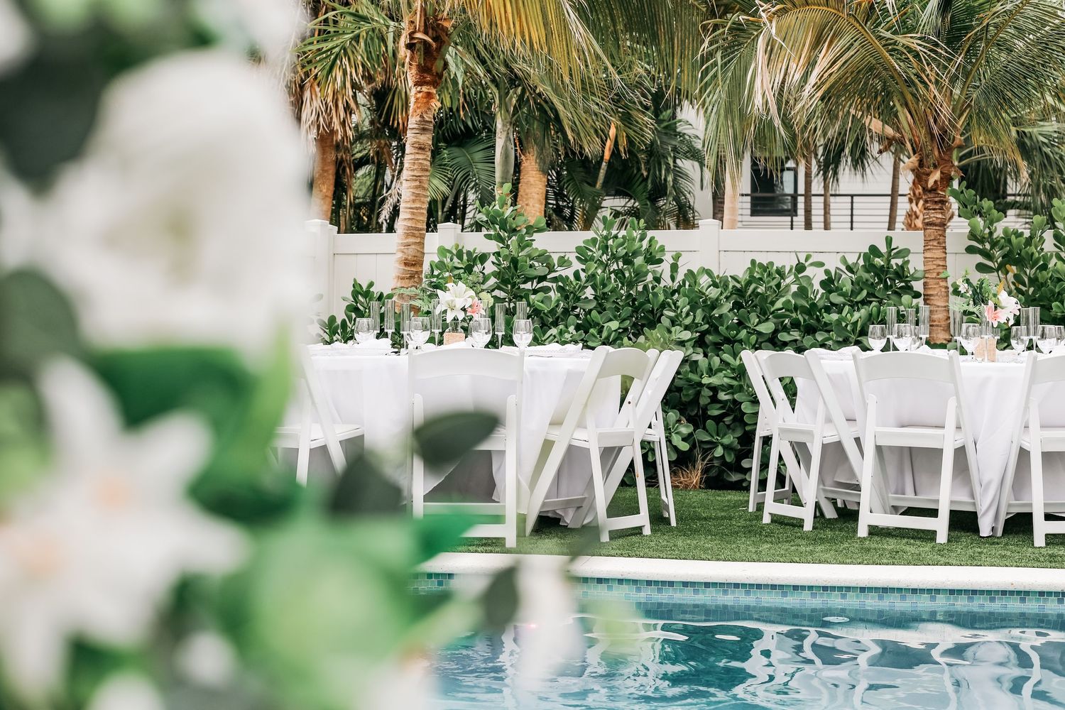 Elegant outdoor poolside wedding reception with white chairs and tables under lush palm trees.