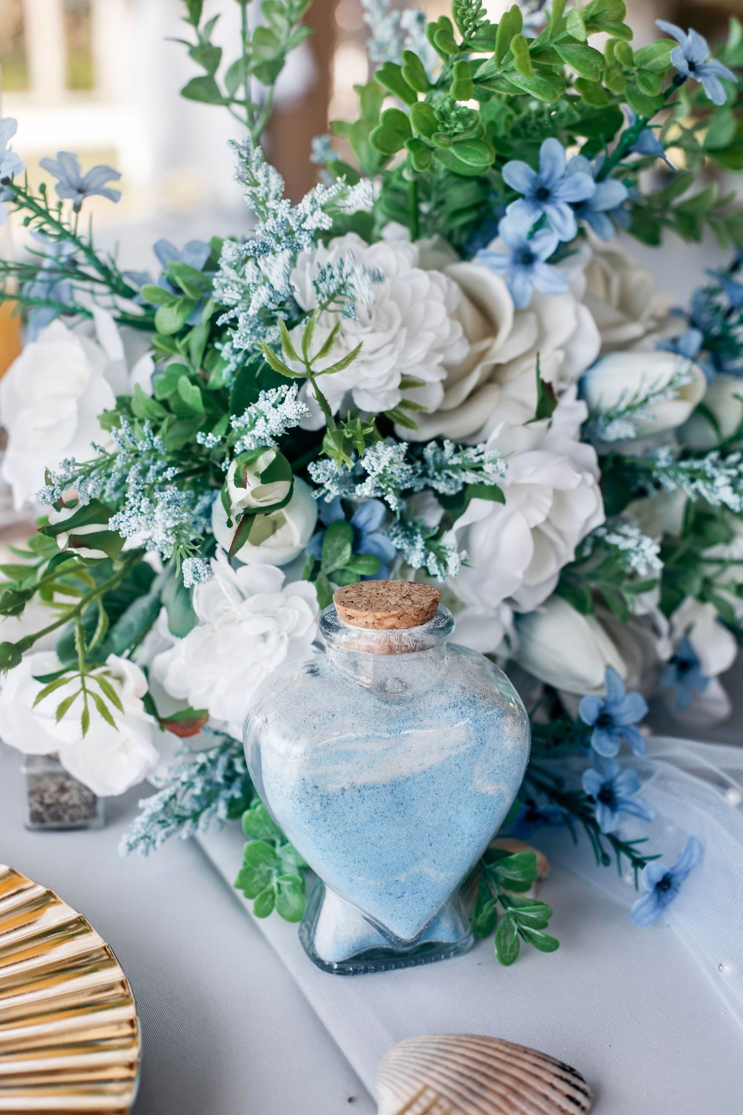 Blue and white floral arrangement with heart-shaped sand bottle and seashells on a white table.