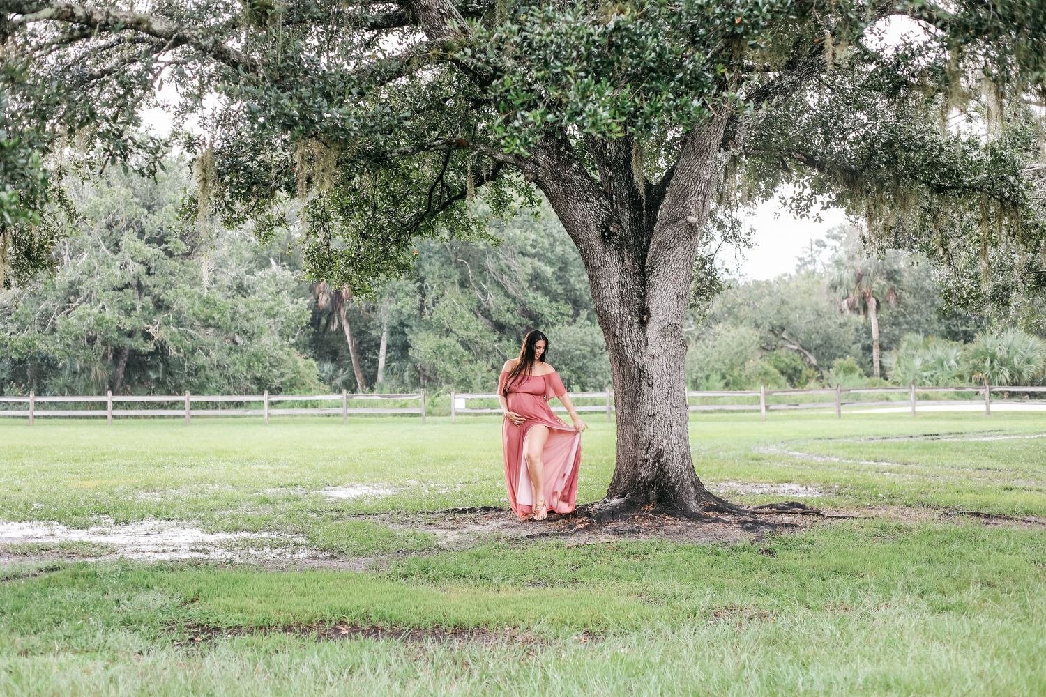 Pregnant woman in coral maxi dress leaning against large oak tree in lush green park setting.