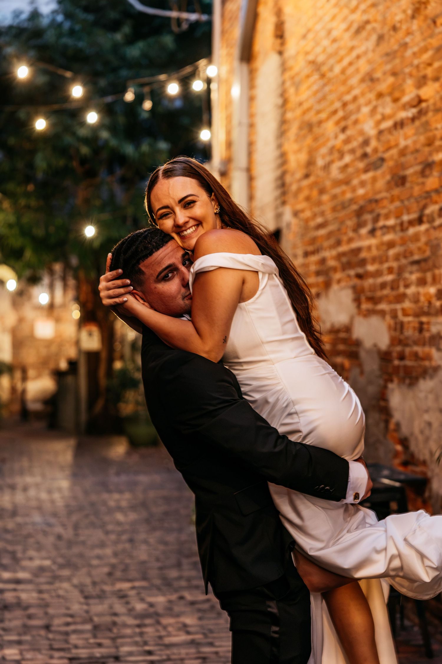 Groom lifts bride in white off-shoulder gown on cobblestone alley with string lights at night.