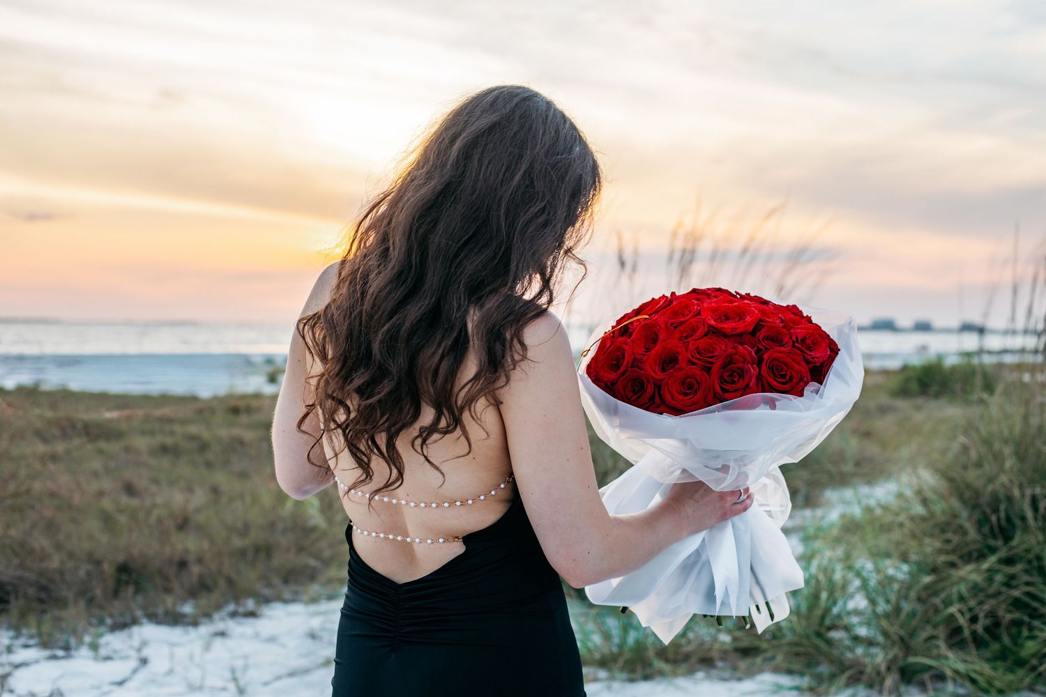 Woman in black dress holding large red rose bouquet on sandy beach at sunset.