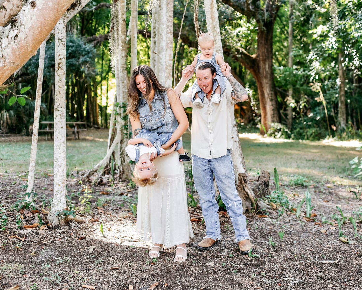 Candid family photoshoot in a natural outdoor setting with trees and dappled sunlight.
