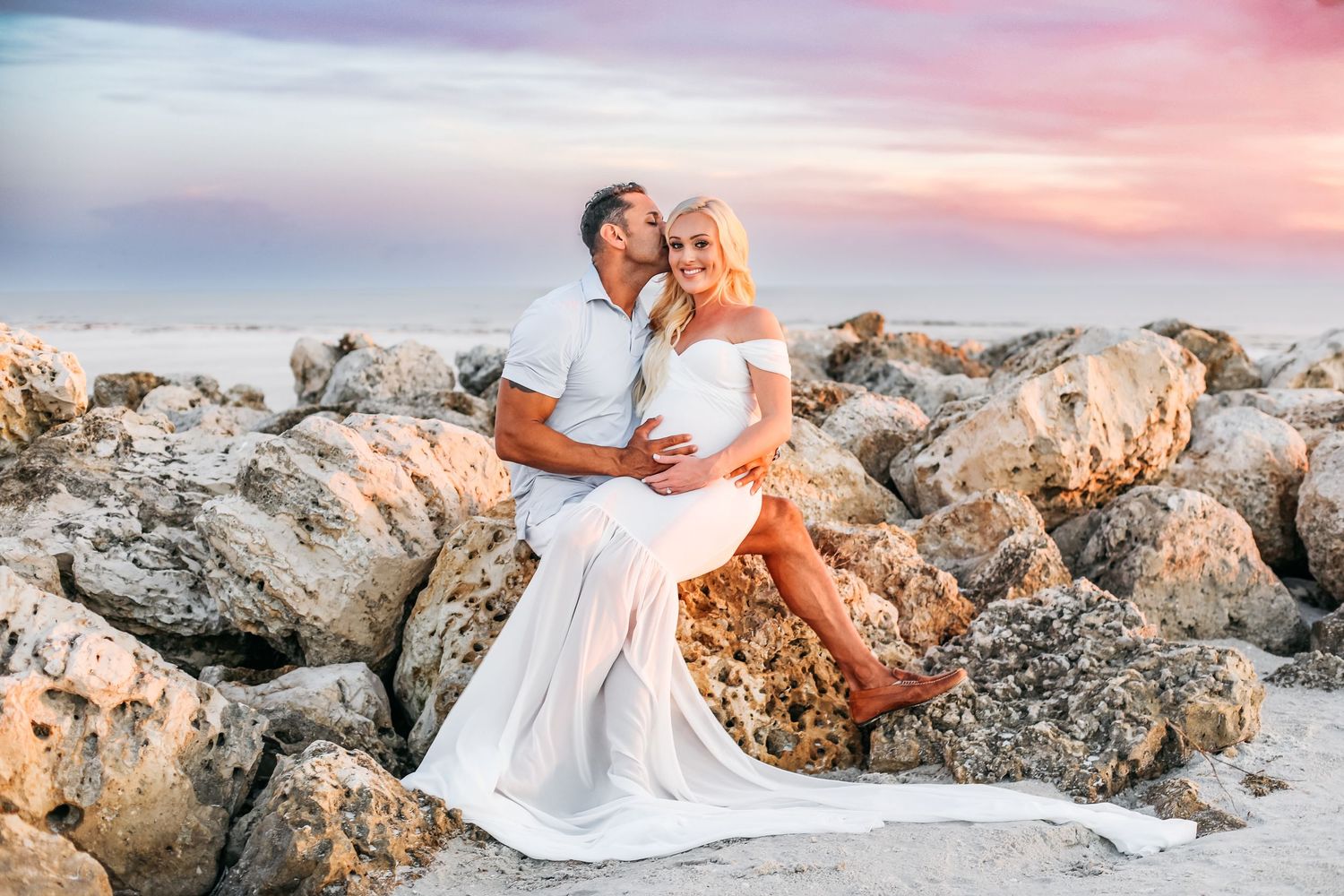 A couple in wedding attire embraces on a rocky beach at sunset with soft pink skies in the background.