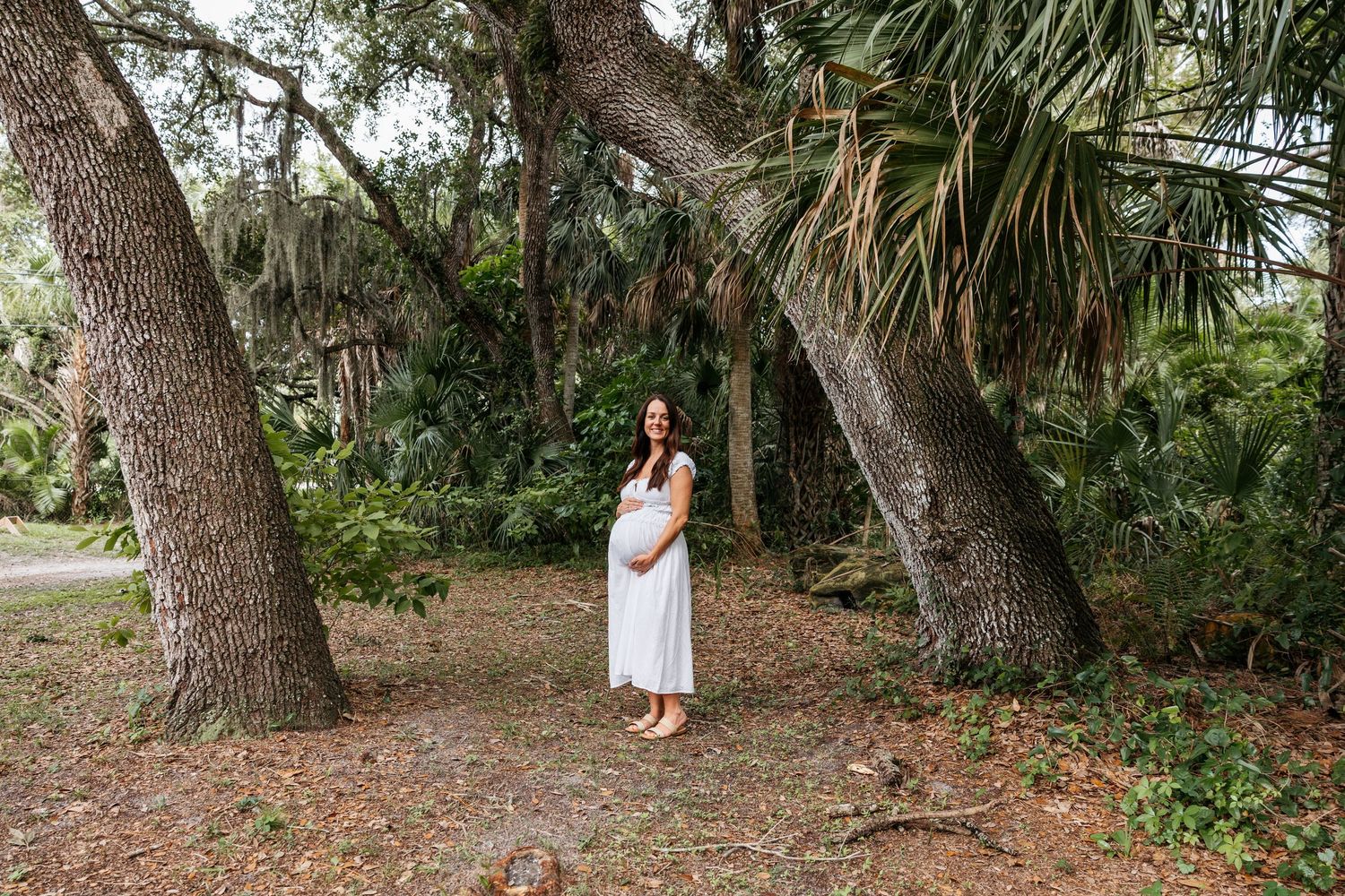 A pregnant woman in a white dress stands peacefully between two large trees in a lush green outdoor setting.