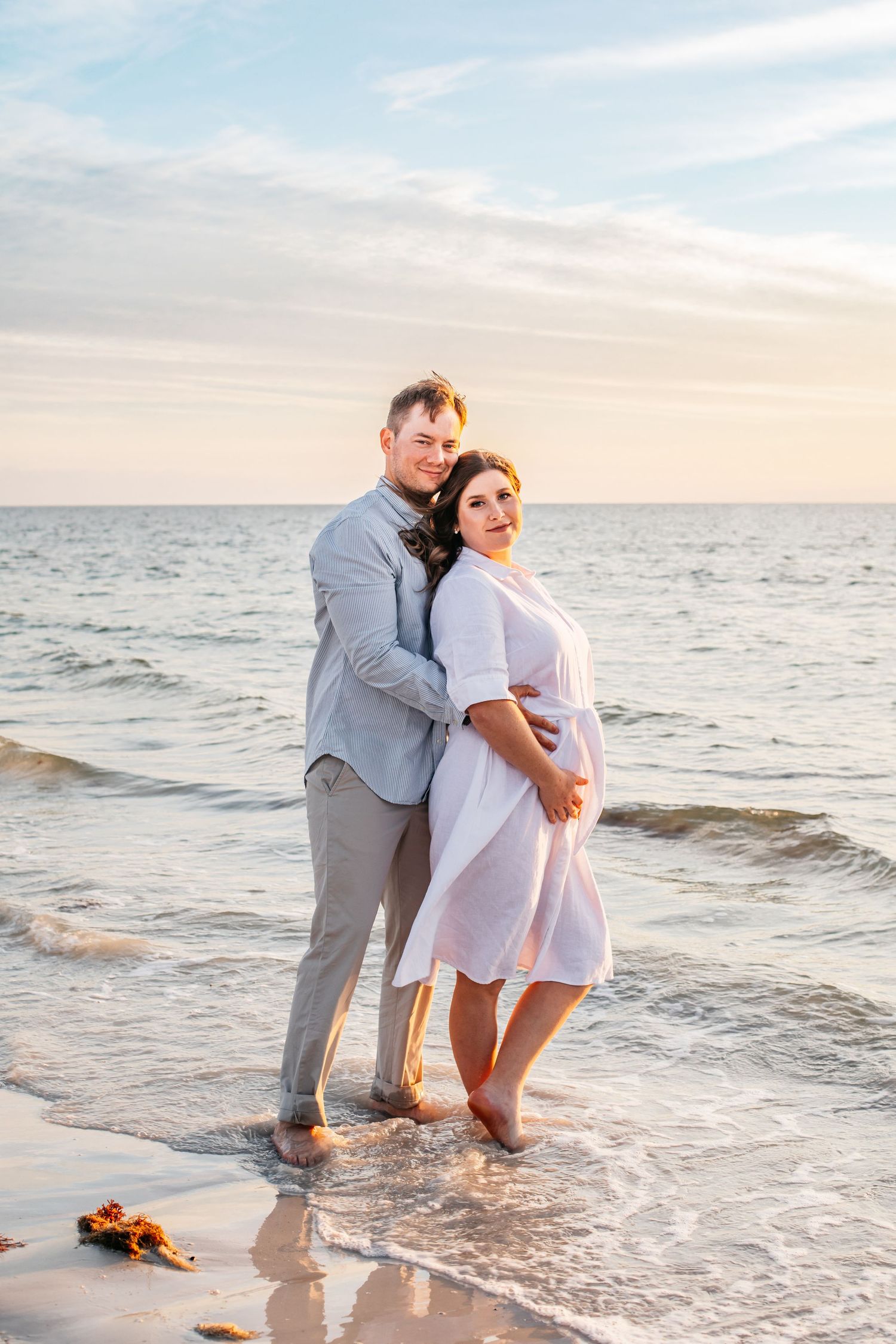 A couple stands barefoot on a sandy beach at sunset, the woman in a white dress cradling her baby bump.