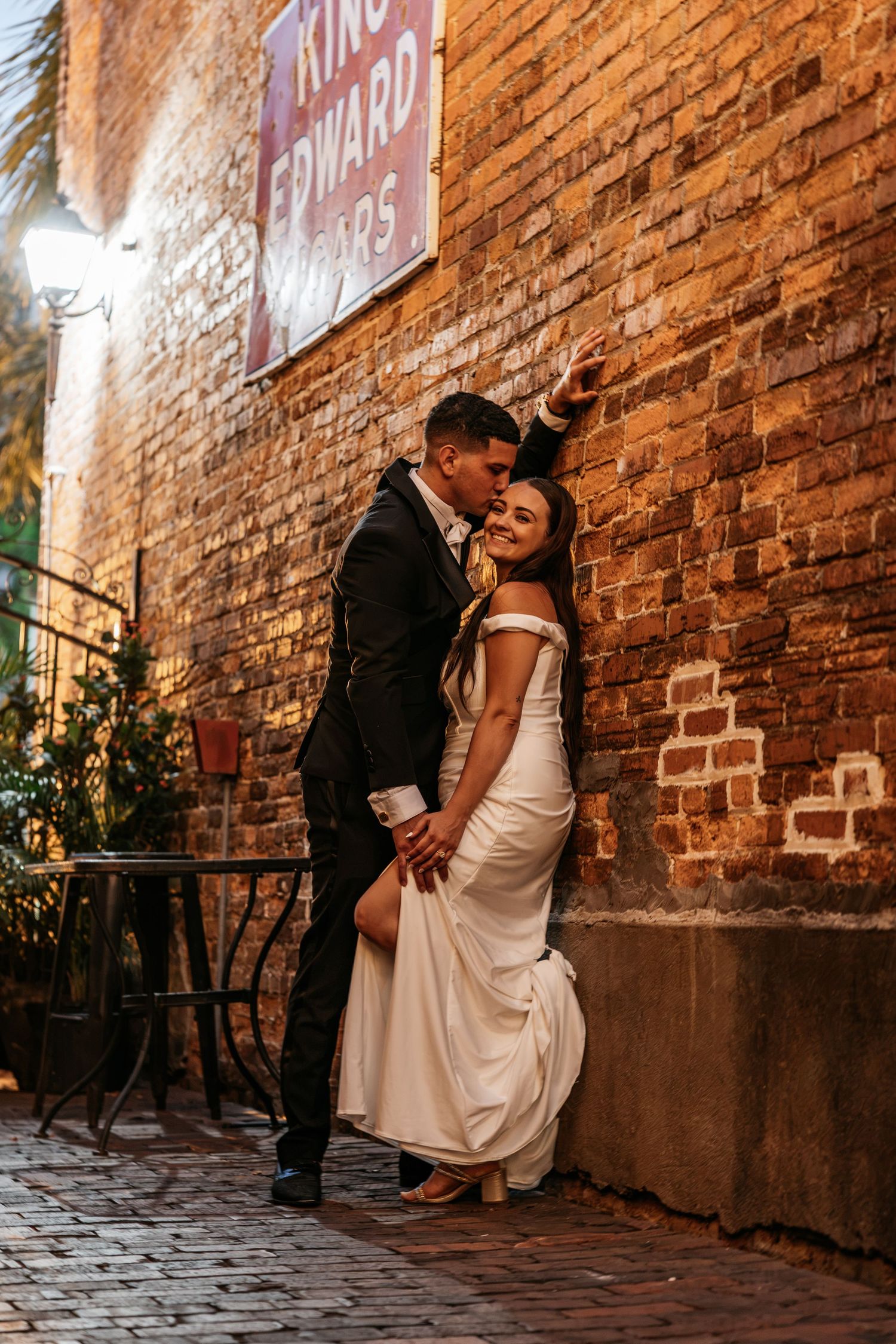 Bride and groom share romantic moment against brick wall on cobblestone street at night.
