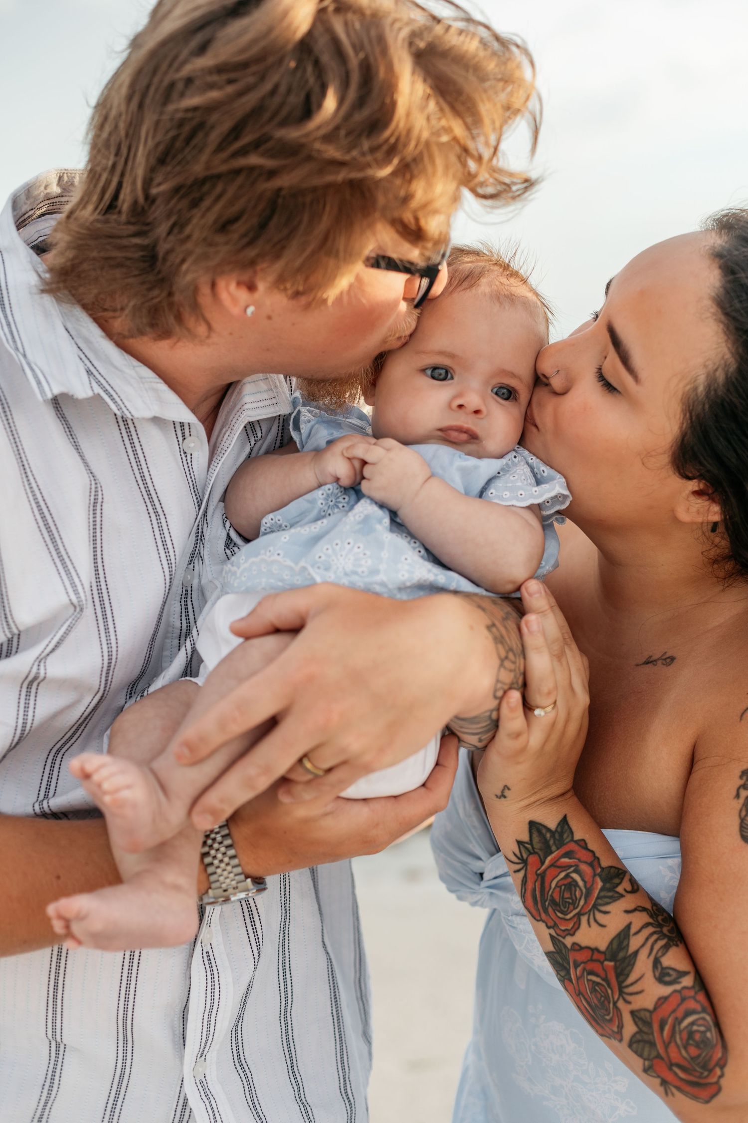 Two parents kissing their baby on the cheeks at the beach, mother with rose tattoos on her arm.