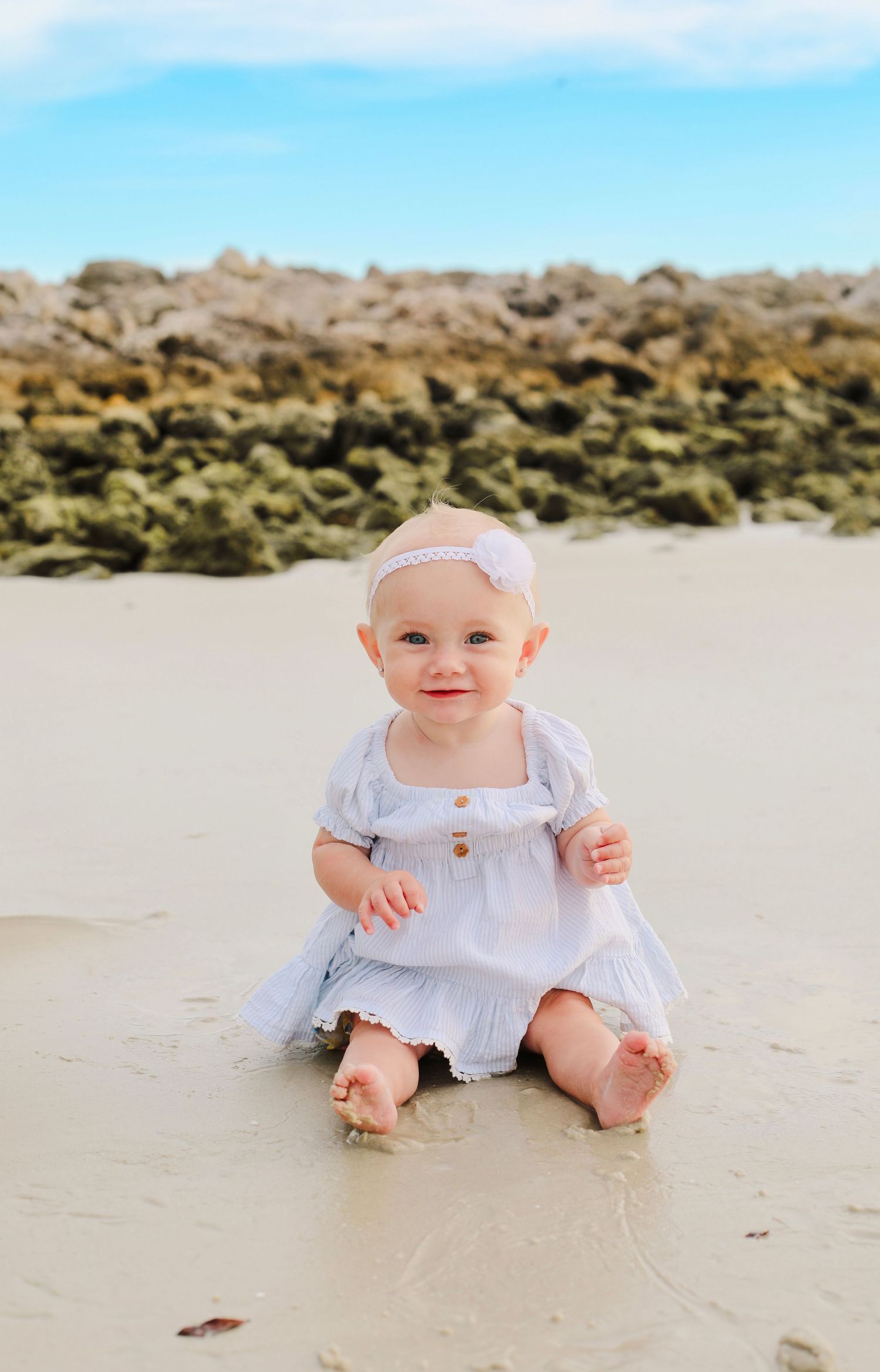 Baby wearing white dress sits on sandy beach with rocky background.