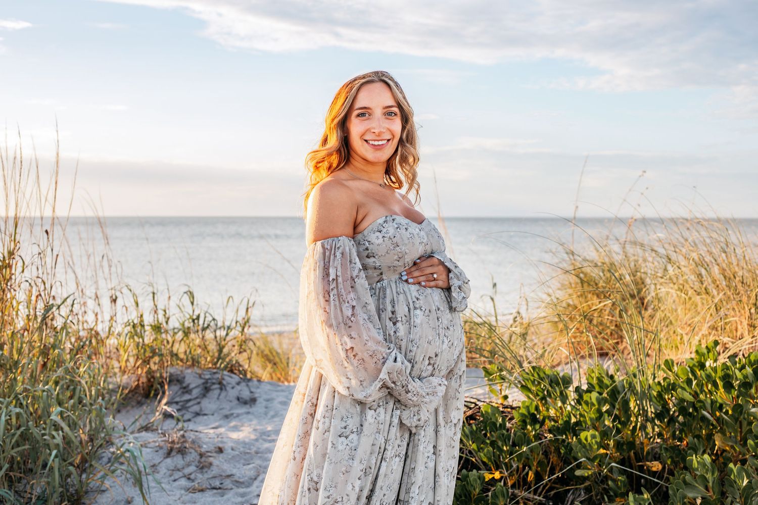 Expectant mother in flowing off-shoulder lace dress poses on beach dunes during golden sunset.