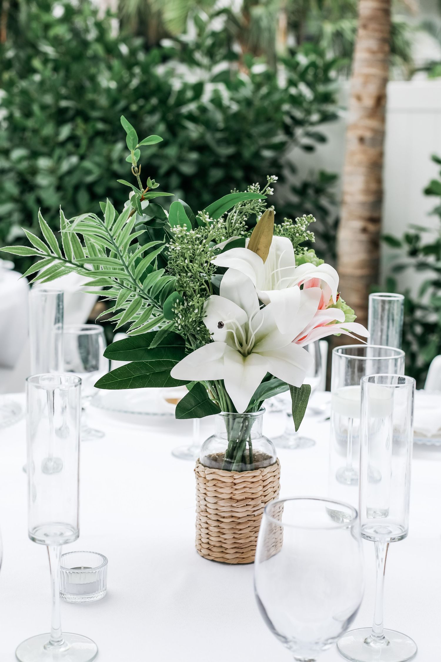 White lilies in a wicker vase centerpiece on a white tablecloth with glass stemware at an outdoor event.