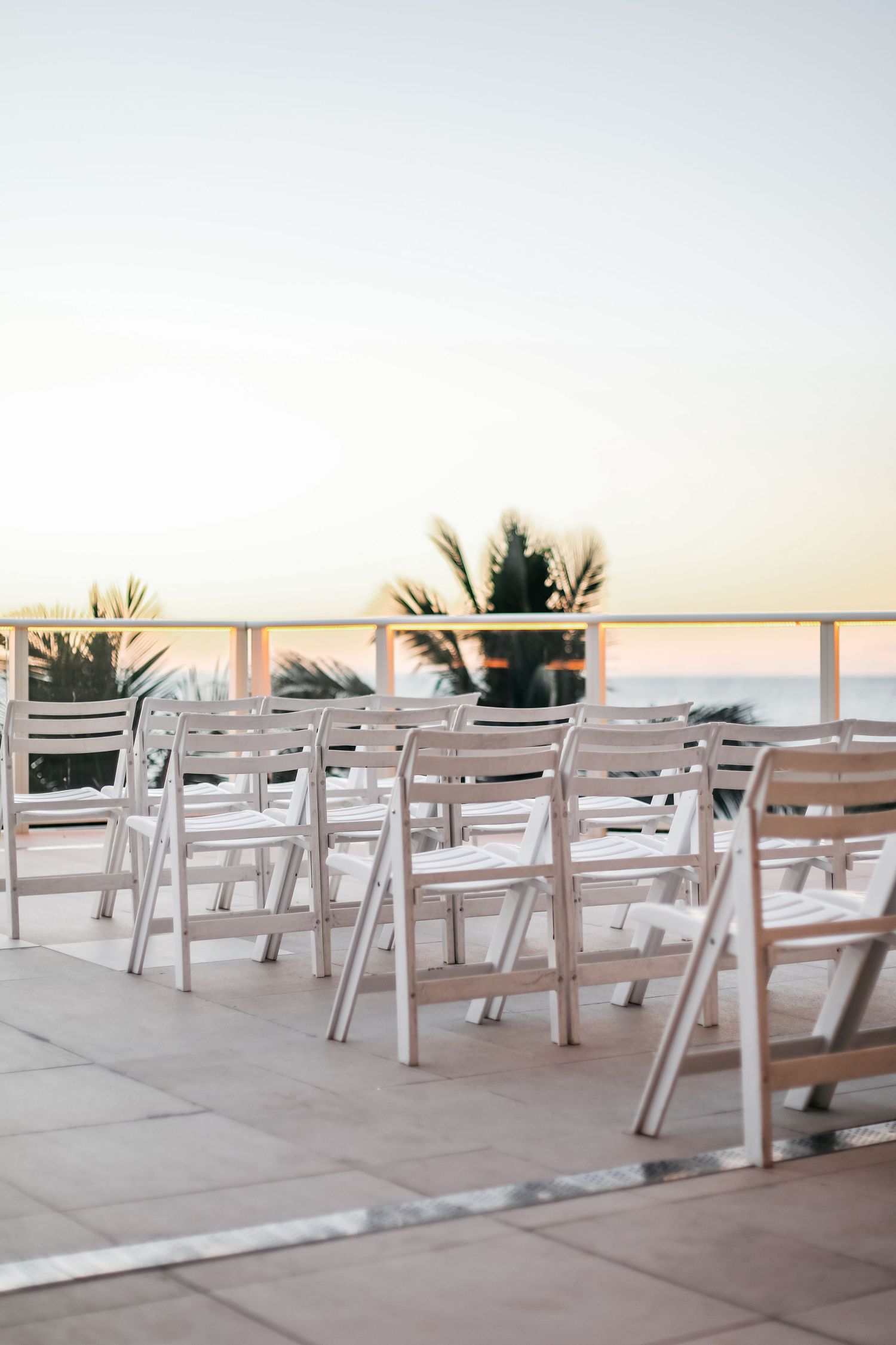 White folding chairs arranged on an outdoor terrace overlooking the ocean at sunset with palm trees in the background.