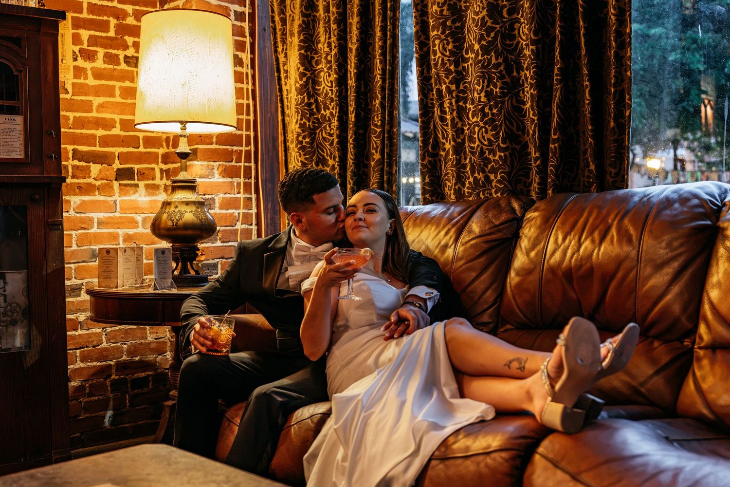 Romantic couple in wedding attire relaxing on a leather sofa in a warmly lit room with brick walls and gold curtains.