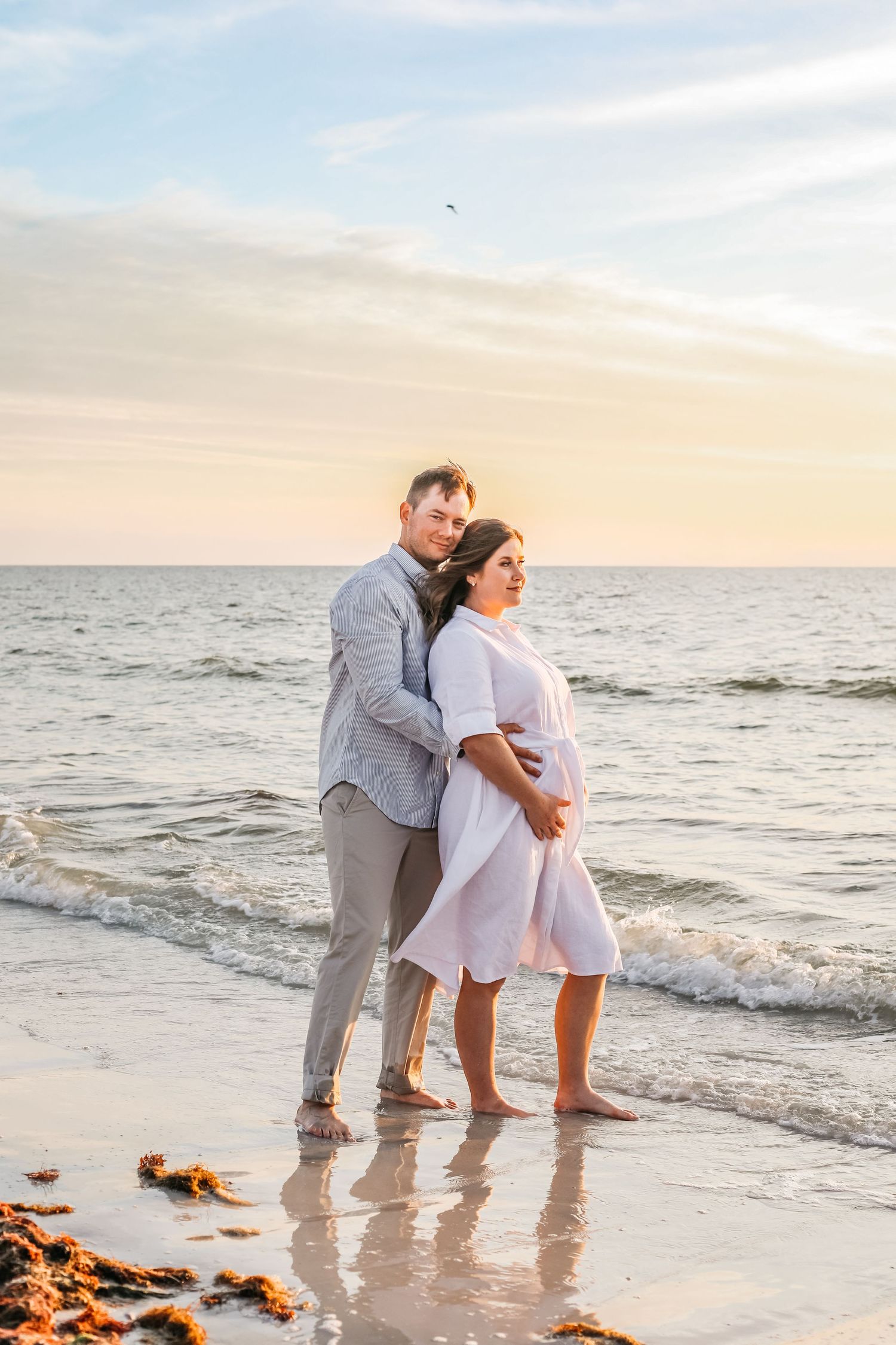 Pregnant couple standing barefoot on a sandy beach at sunset, woman in white dress with hands on belly.