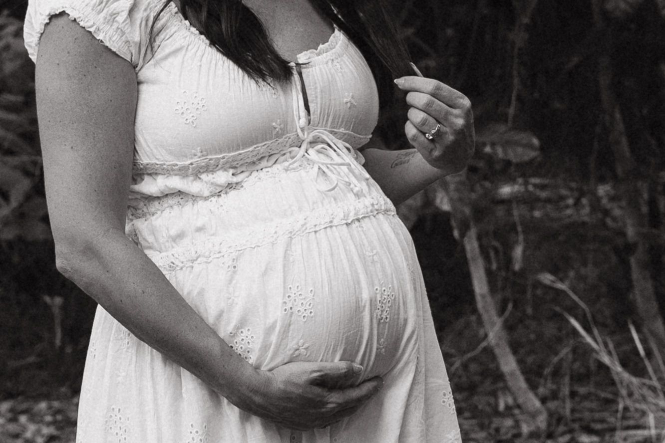Black and white photo of a pregnant woman in a white dress cradling her baby bump outdoors.