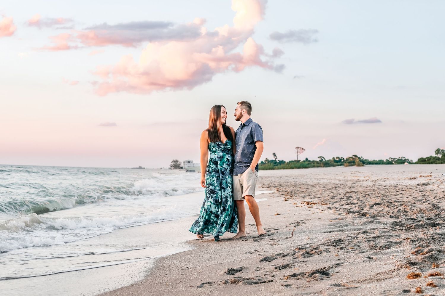 A couple walks along a beach at sunset with waves crashing nearby as pink clouds fill the sky.