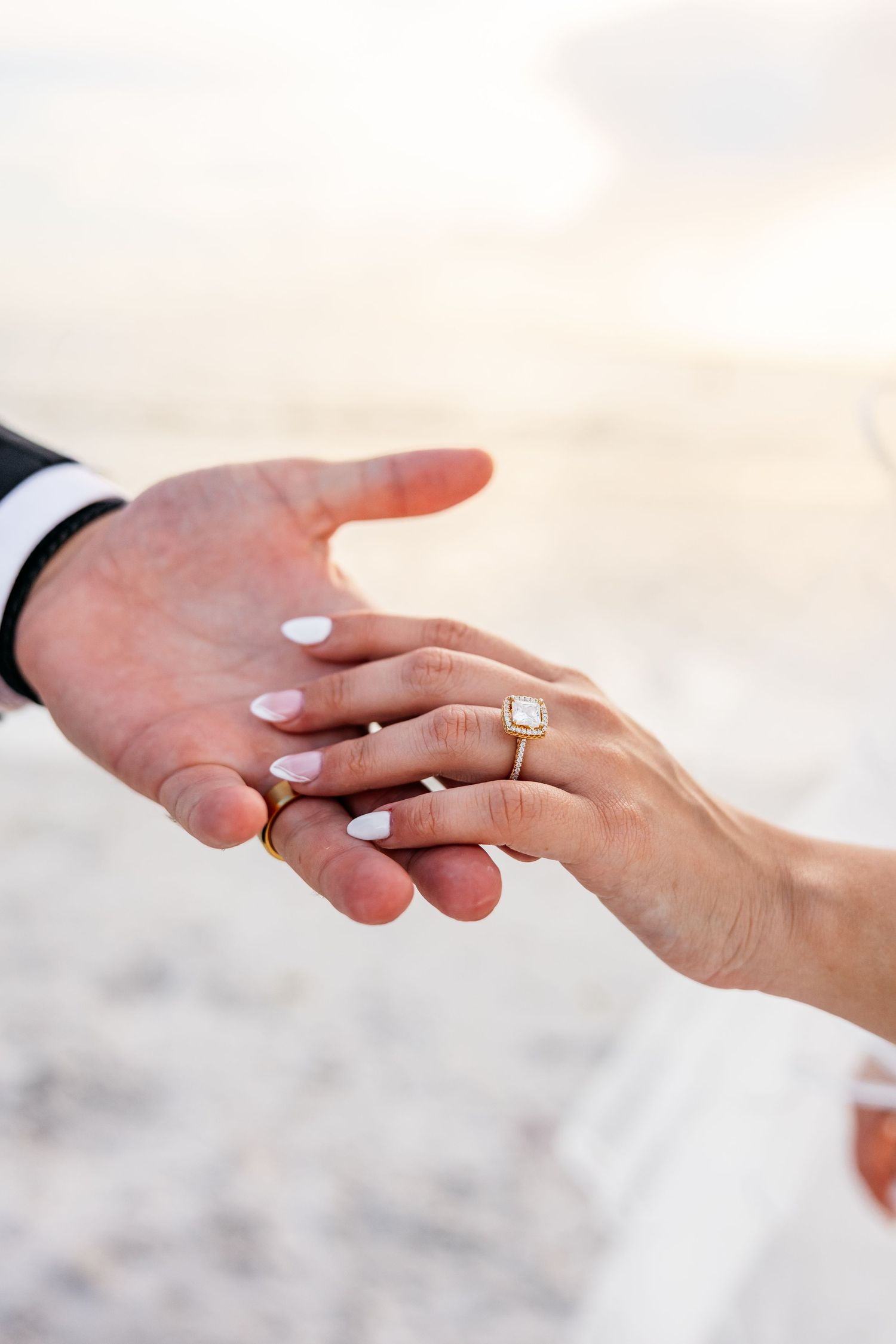 Close-up of a couple holding hands on a beach, woman wearing a gold diamond engagement ring with white manicured nails.