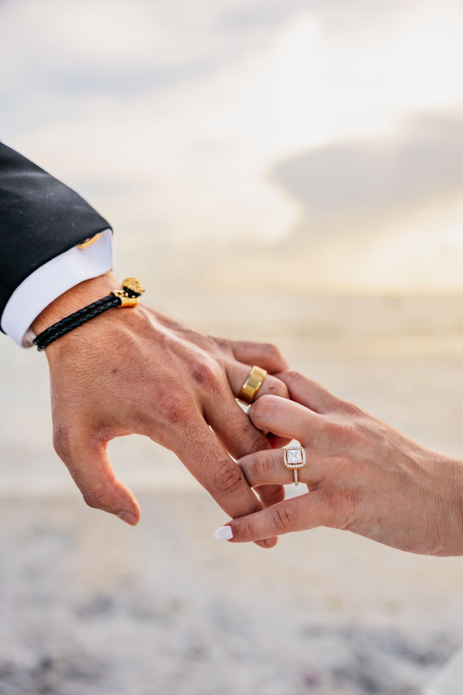 Close-up of newlywed couple holding hands on beach, showing diamond engagement ring and gold wedding bands.