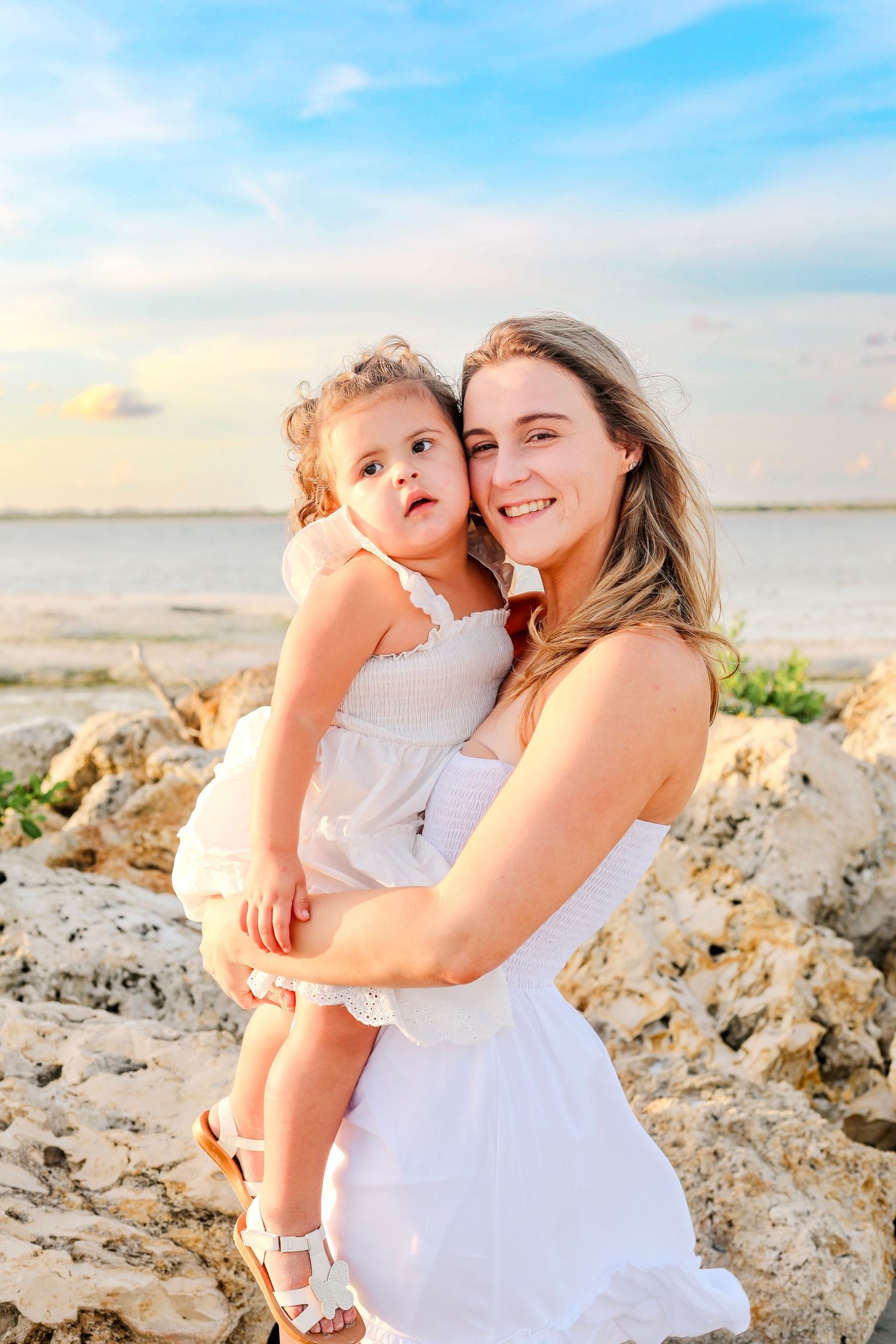 Parent and child wearing white dresses embrace at sunset on a rocky beach with blue sky and ocean in background.