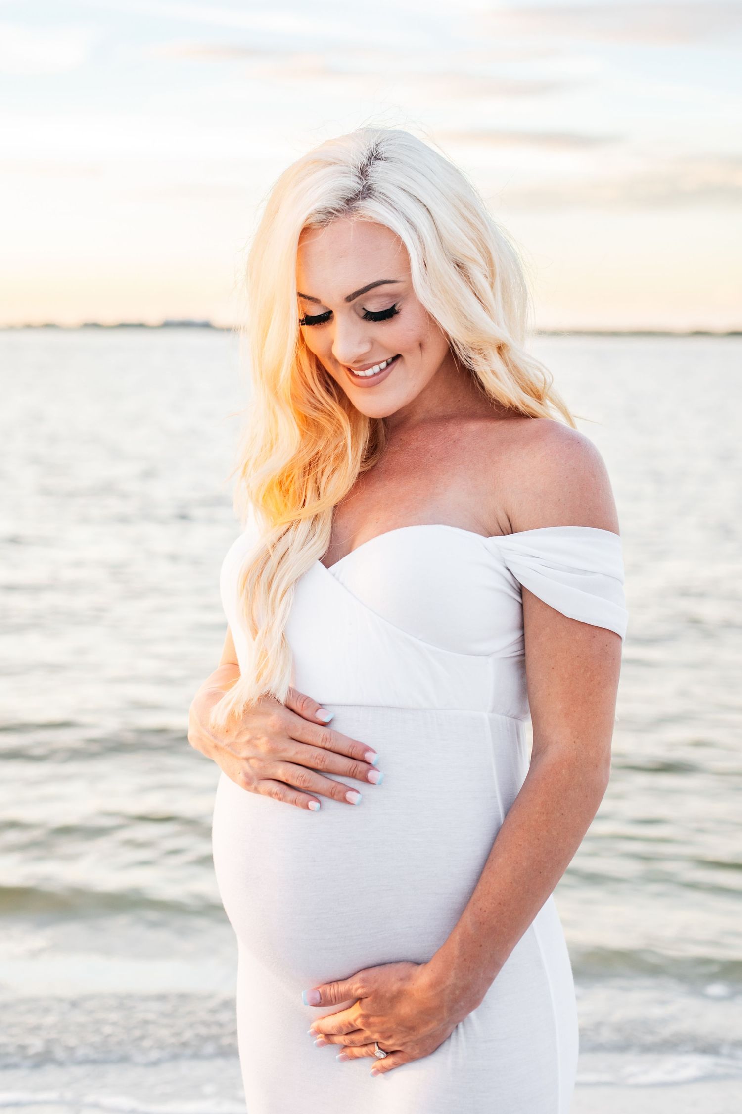Expectant mother in white dress caressing baby bump on beach at sunset.