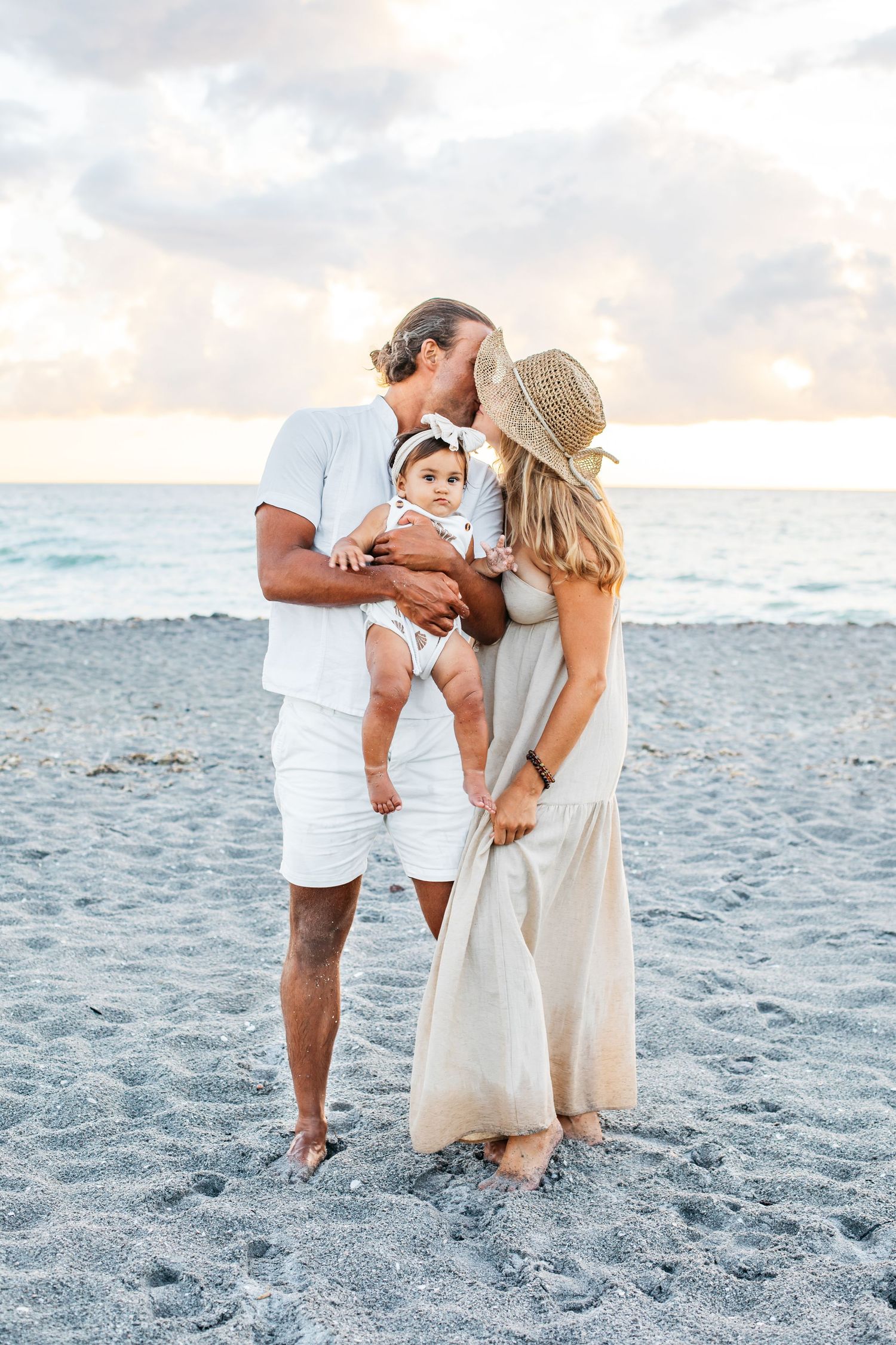 A family shares tender moments together on a sandy beach at sunset wearing coordinated white and neutral outfits.
