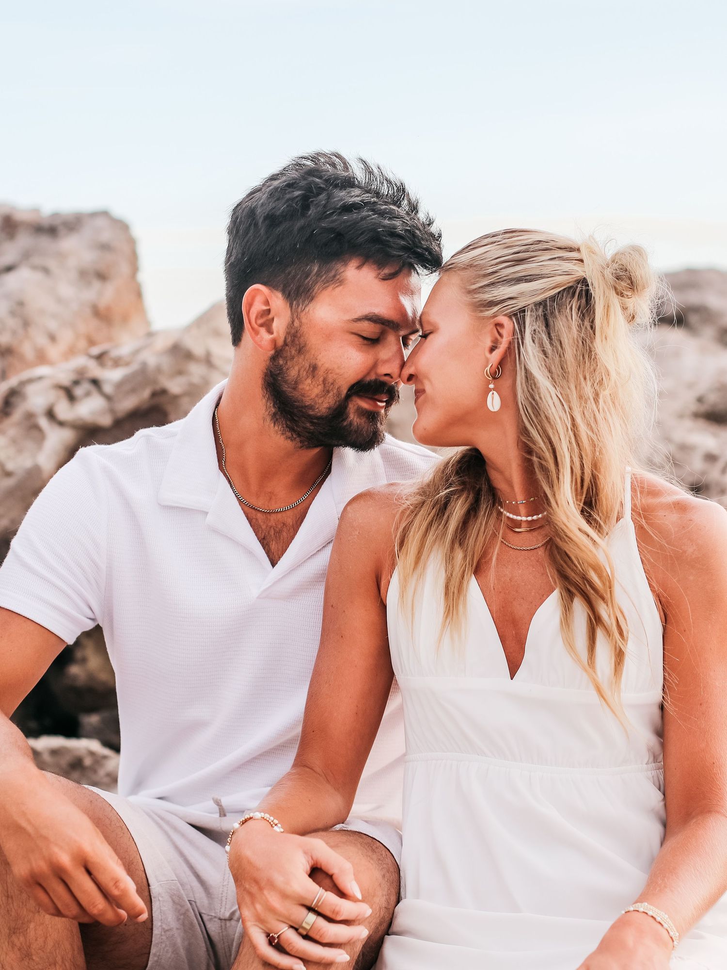 Couple in white outfits embracing on beach rocks during golden hour photoshoot.