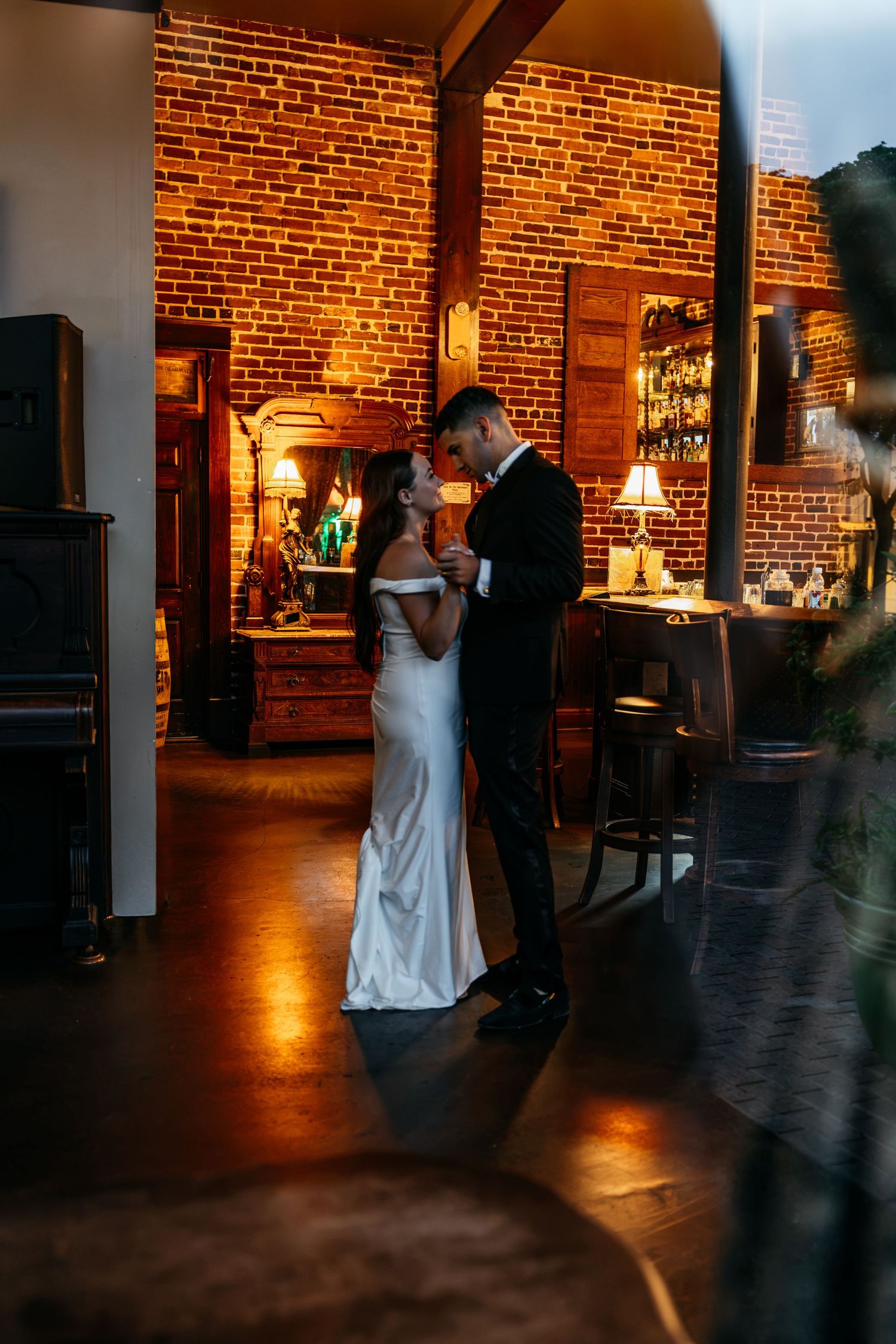 Bride and groom share a romantic first dance in a warmly lit venue with exposed brick walls.