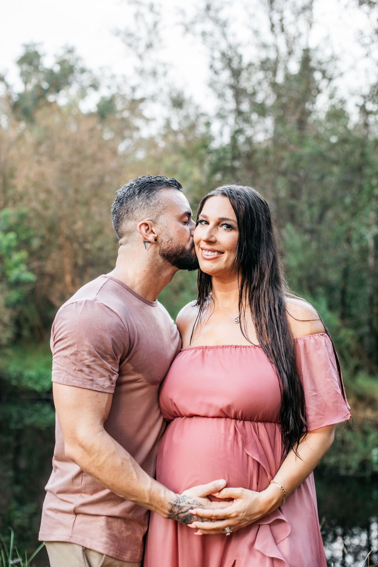 A couple in pink and mauve outfits share intimate moments during a maternity photoshoot in a natural outdoor setting.