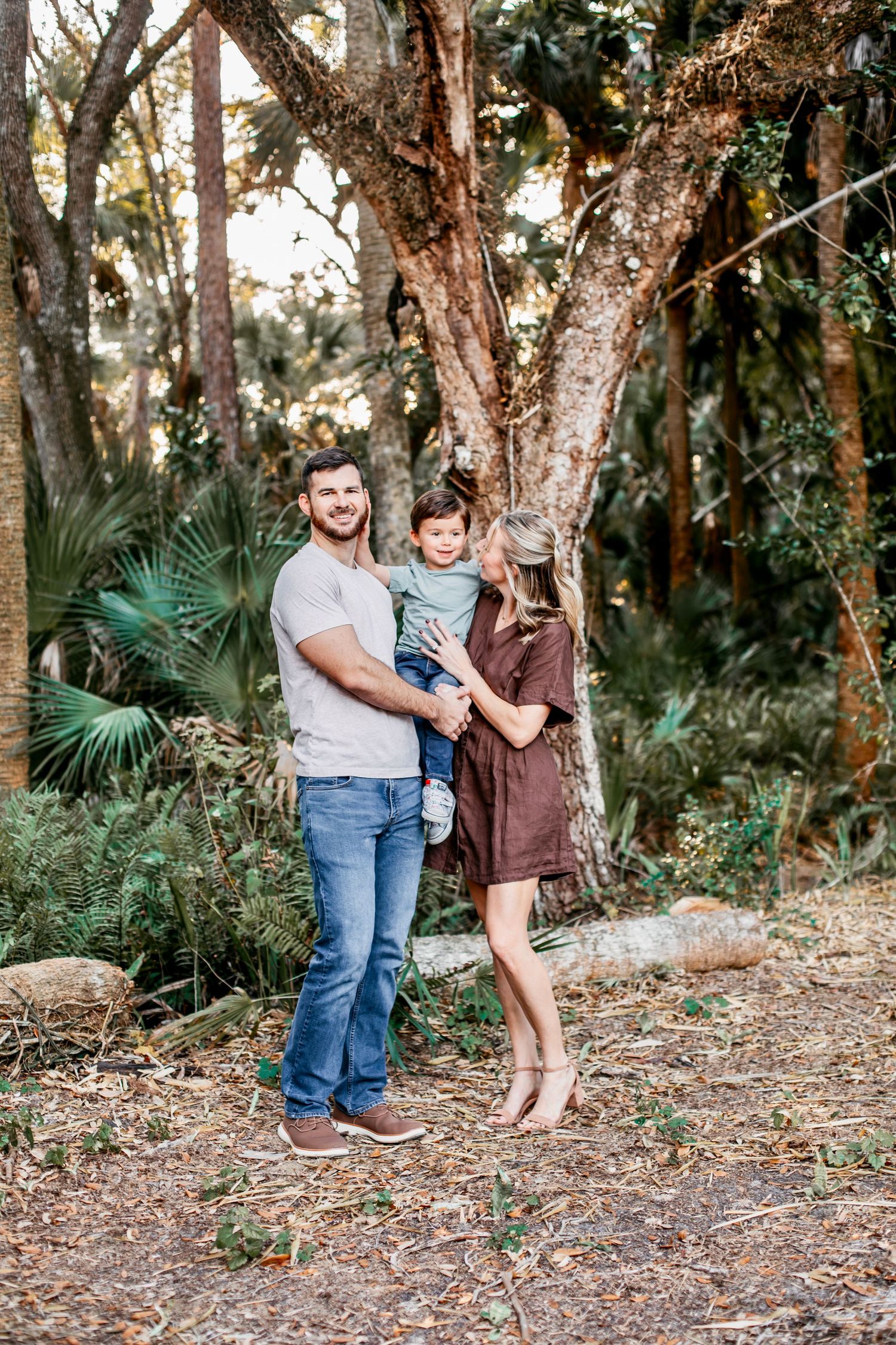 A family enjoys a playful moment together in a wooded outdoor setting with palm trees and natural surroundings.