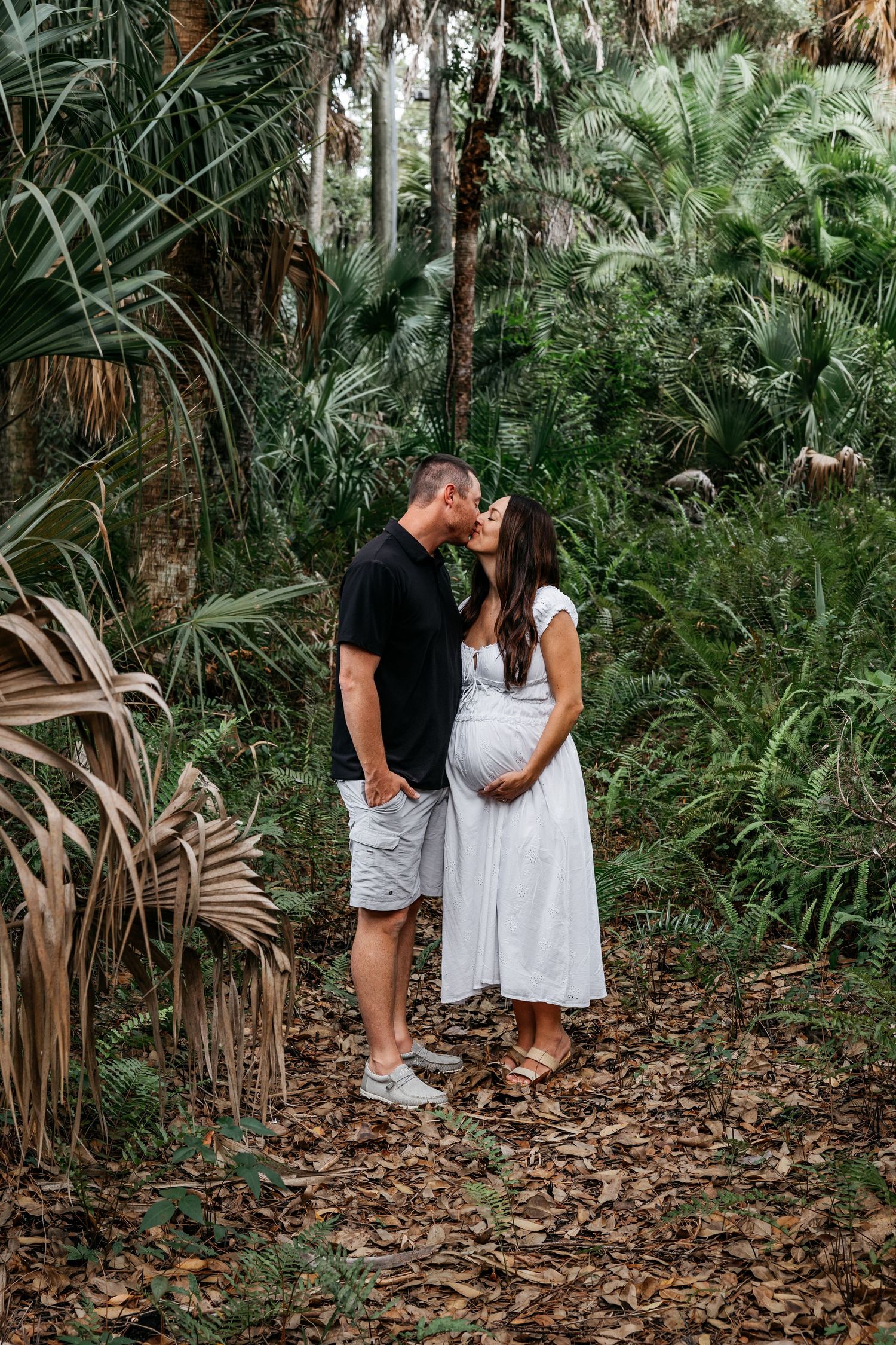 Pregnant couple kissing in a lush tropical forest, woman in white dress cradling baby bump, man in black shirt and grey shorts.