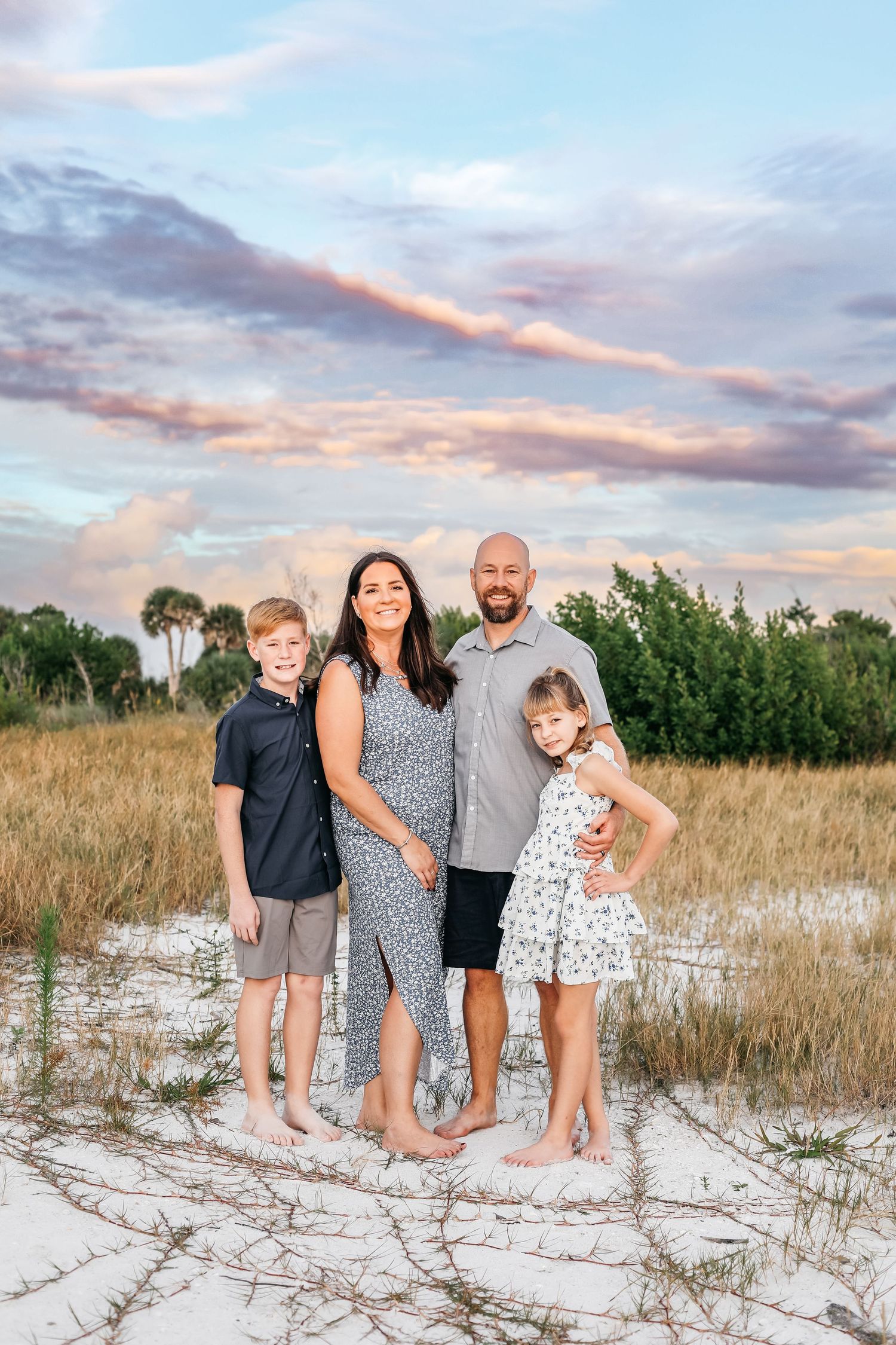 A group stands together on a white sandy beach at sunset with palm trees and tall grass in the background.