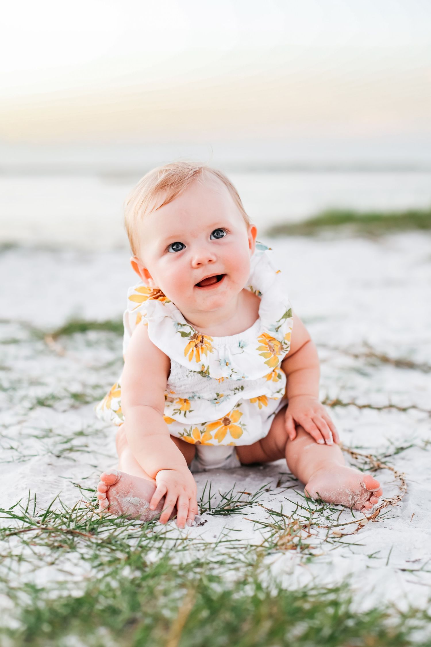 A baby in a floral romper sits on sandy beach during sunset.