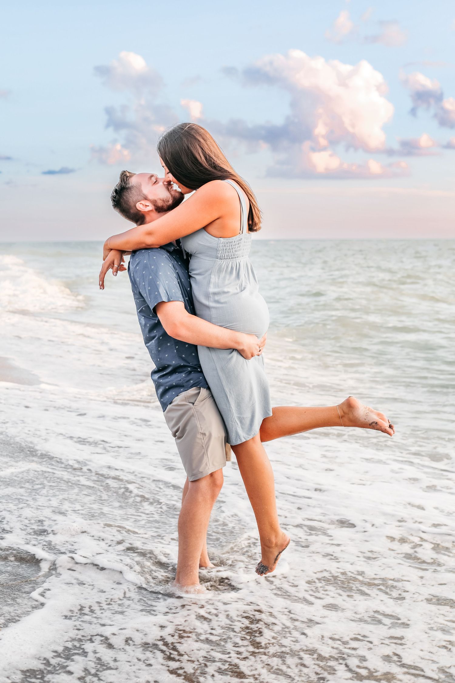 A couple embraces playfully on a beach at sunset with soft waves and pink clouds in the background.