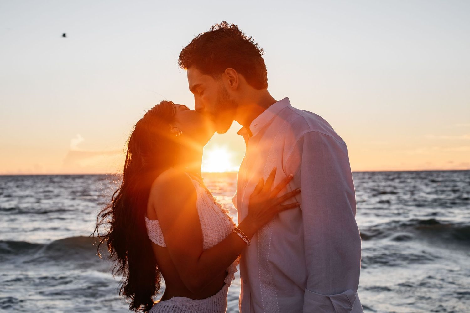 Silhouetted couple embracing on beach at sunset with golden sun flare and ocean waves in background.