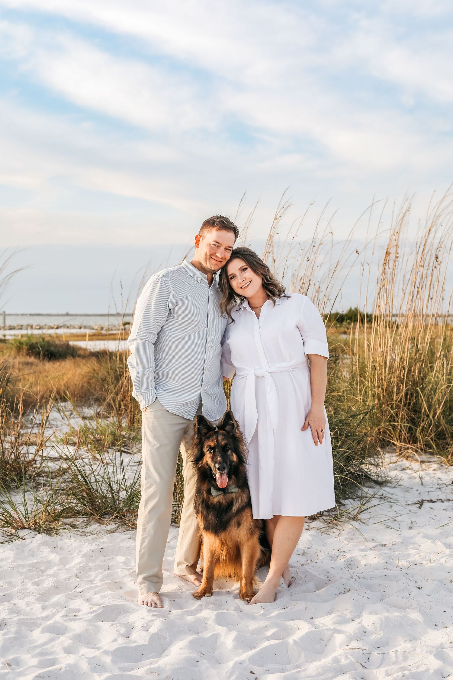 Couple and German Shepherd dog posing on white sand beach with sea oats and blue sky backdrop.