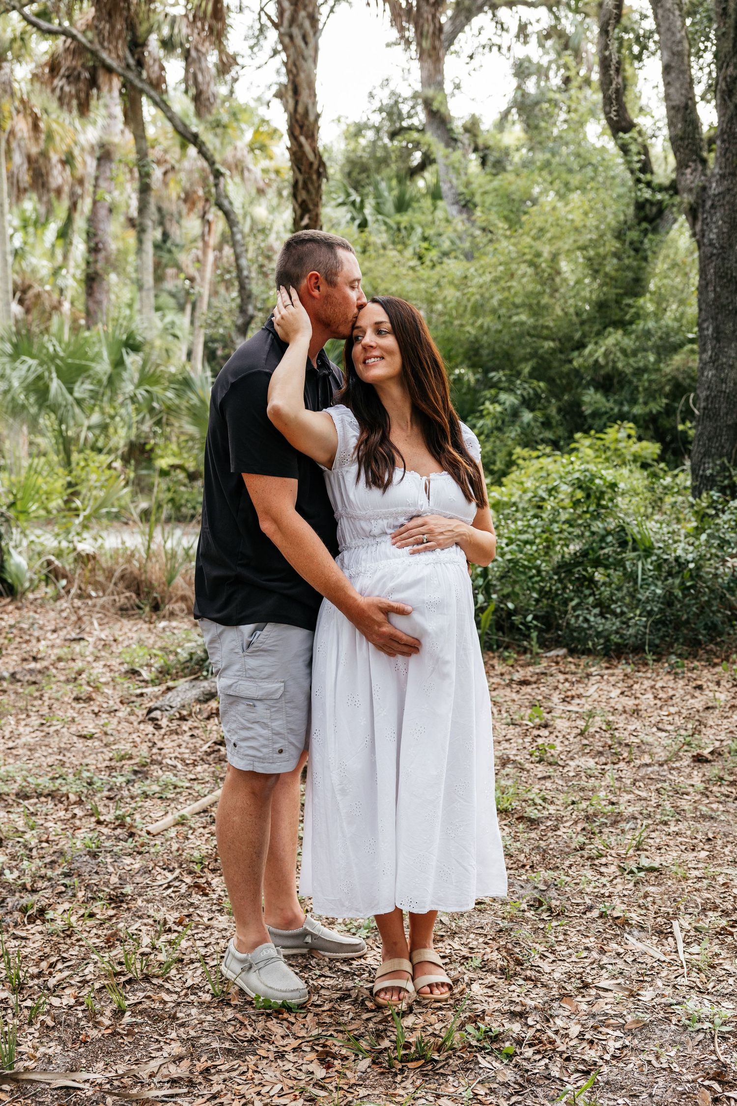 A man kisses a pregnant woman in a white dress outdoors, surrounded by lush green trees in a serene forest setting.