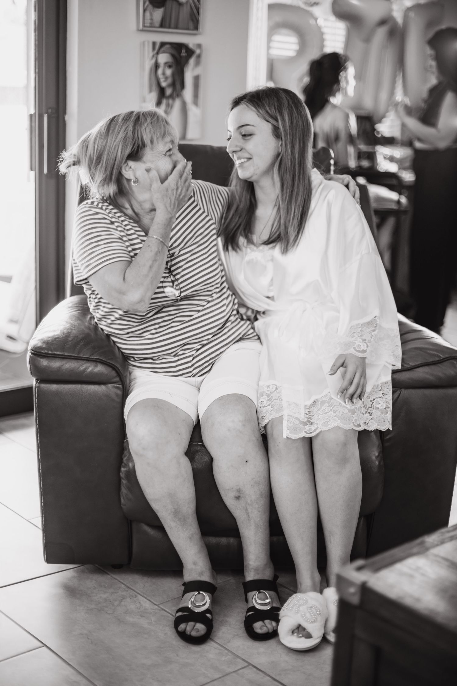 Black and white photo of grandmother and bride sharing a tender moment on a couch during wedding preparations.