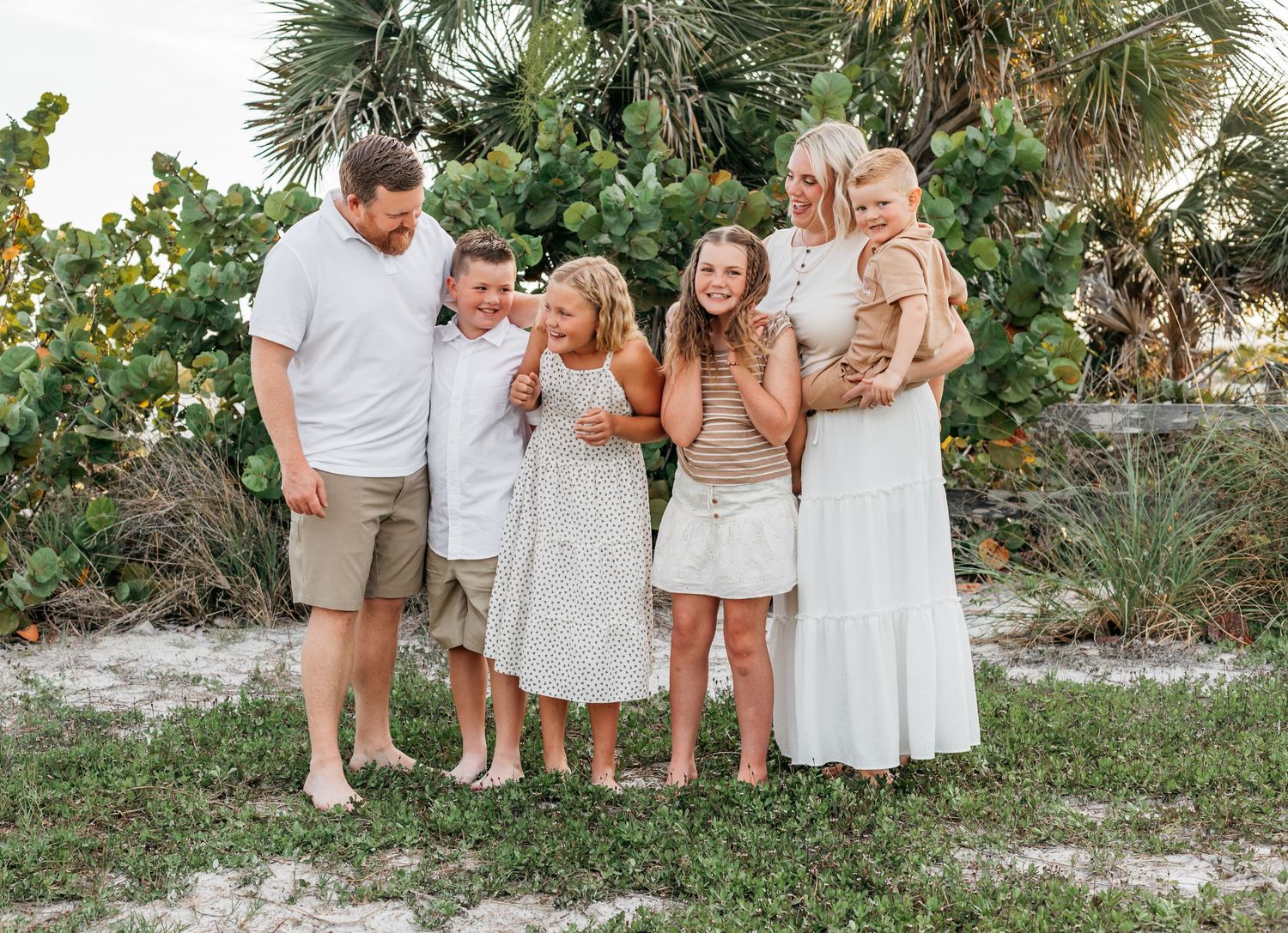 Happy family of six posing together on a sandy beach surrounded by lush tropical greenery.