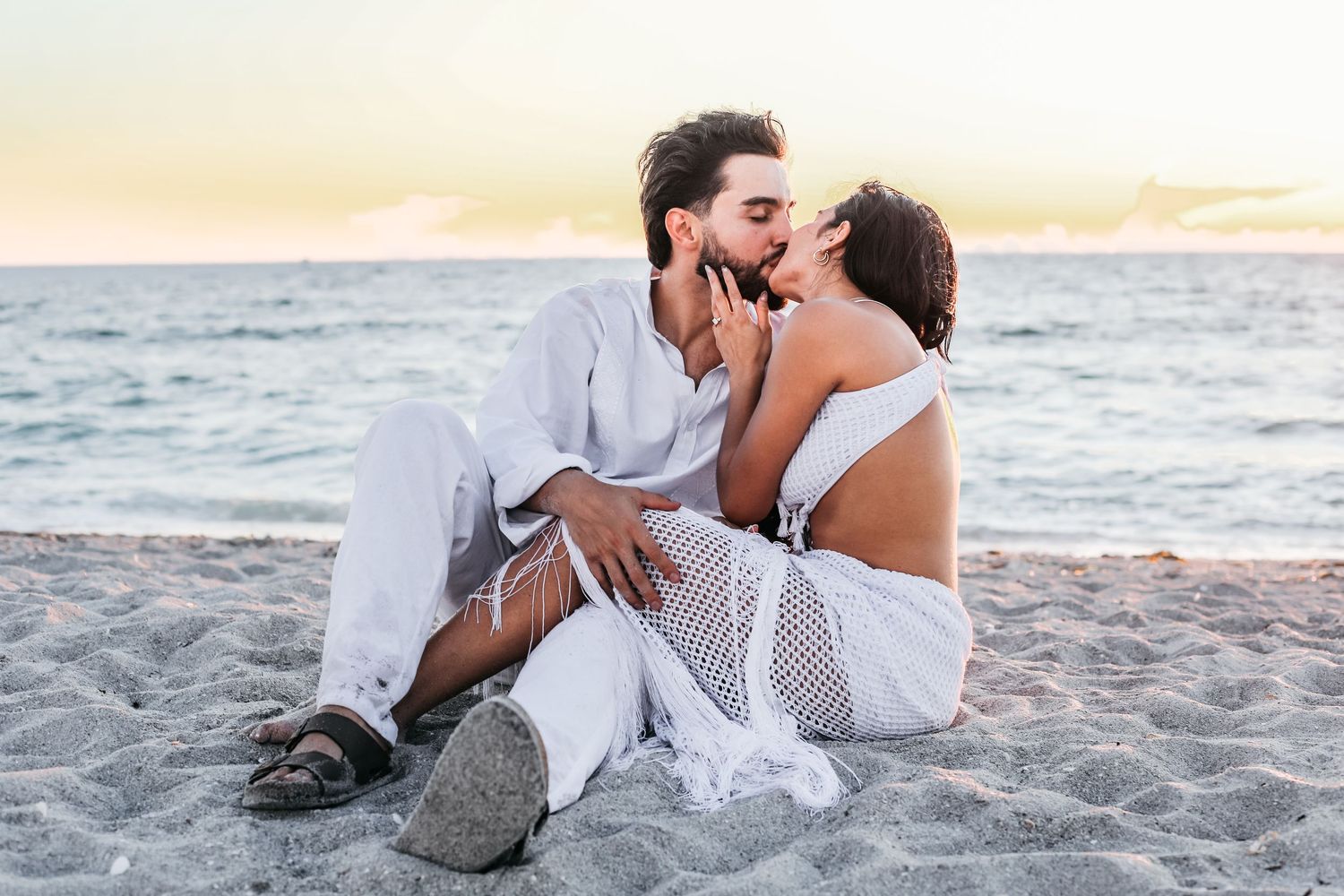 A romantic couple in white outfits share intimate moments while sitting on a sandy beach at sunset.