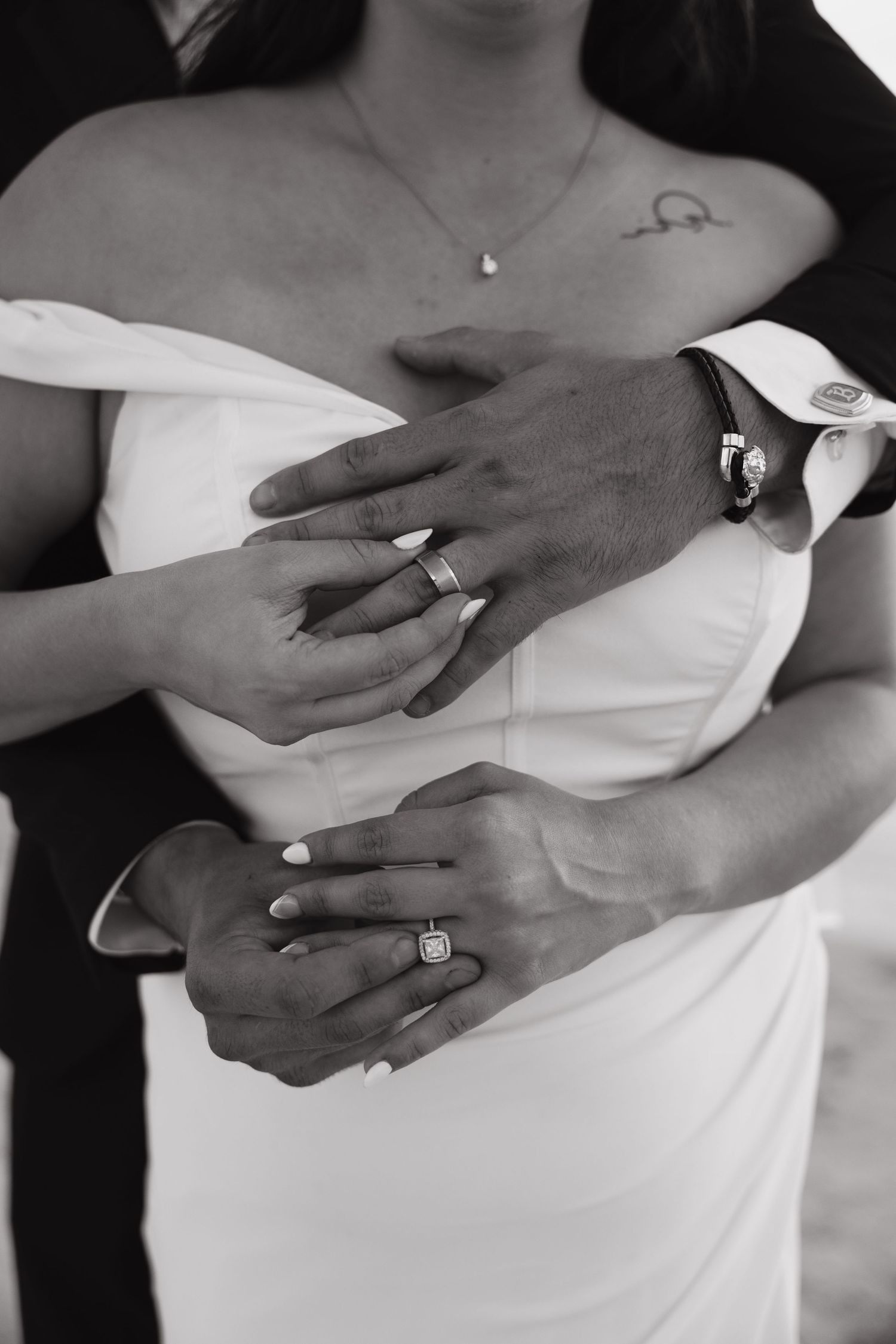 Black and white photo of bride and groom hands intertwined showing wedding rings, bride in white strapless gown.
