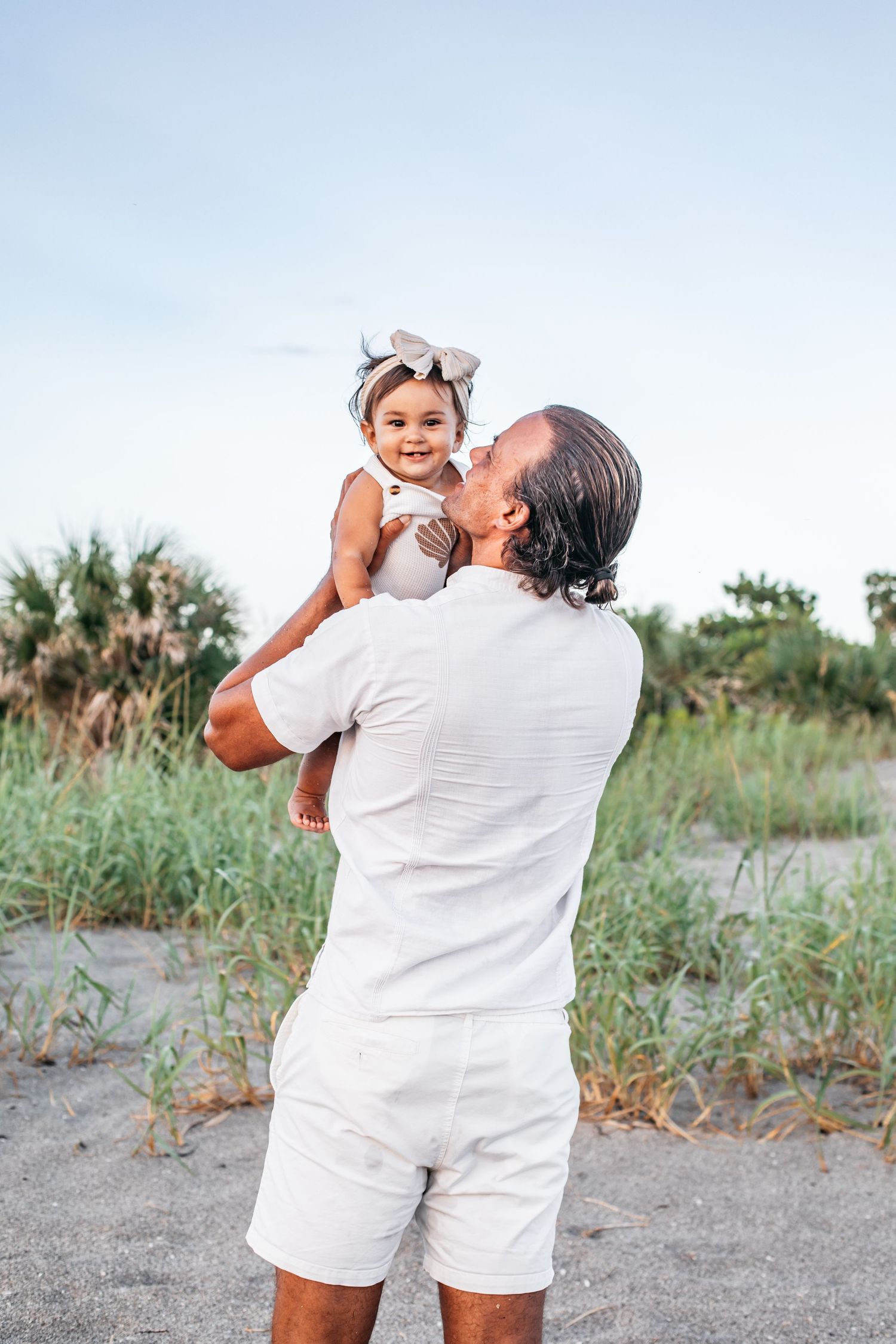 A heartwarming sequence of photos showing a parent in white clothing lifting and playing with a baby on a grassy beach path.