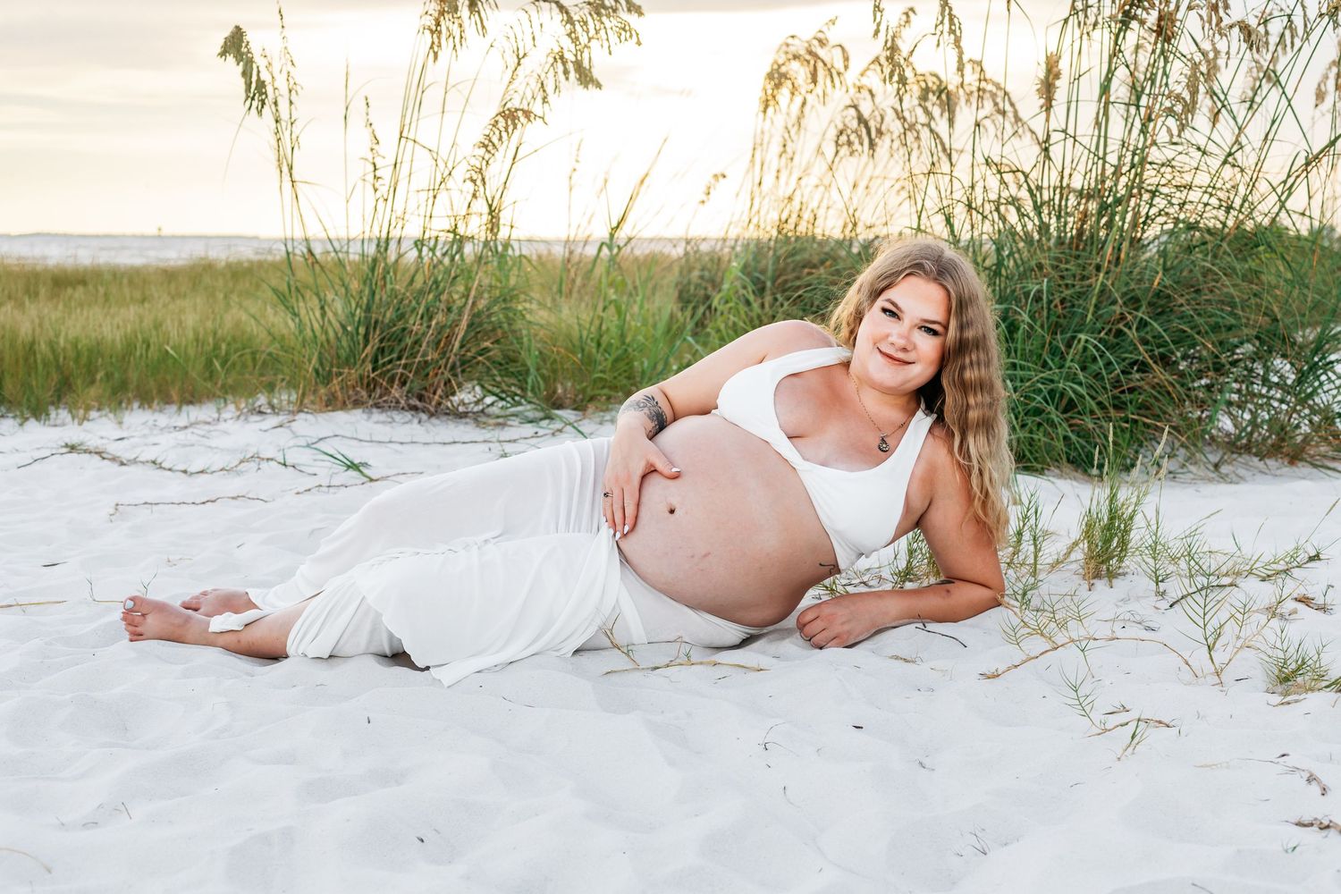Woman in flowing white dress resting on white sand beach with sea oats and ocean in background.