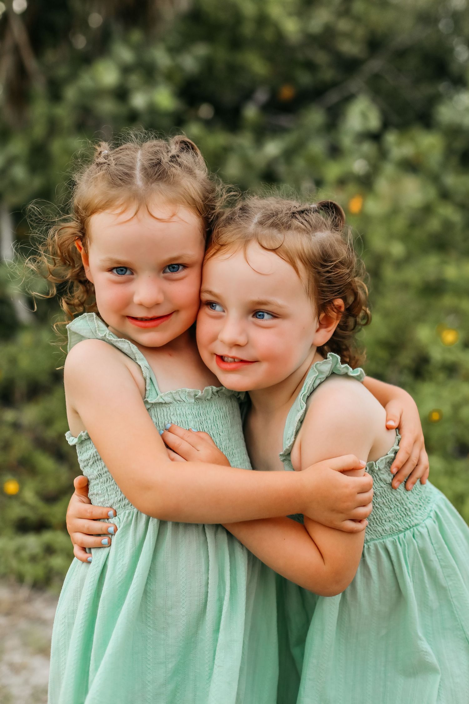 Two young girls in matching mint green dresses hugging outdoors, smiling with curly hair against a green nature background.