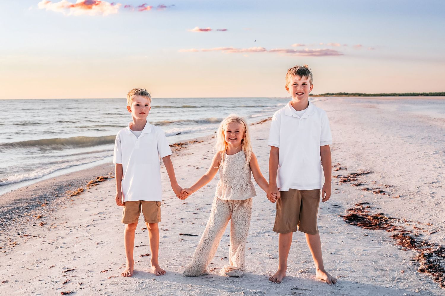 Three siblings in white shirts hold hands while standing on a sunset beach with waves and sand in the background.