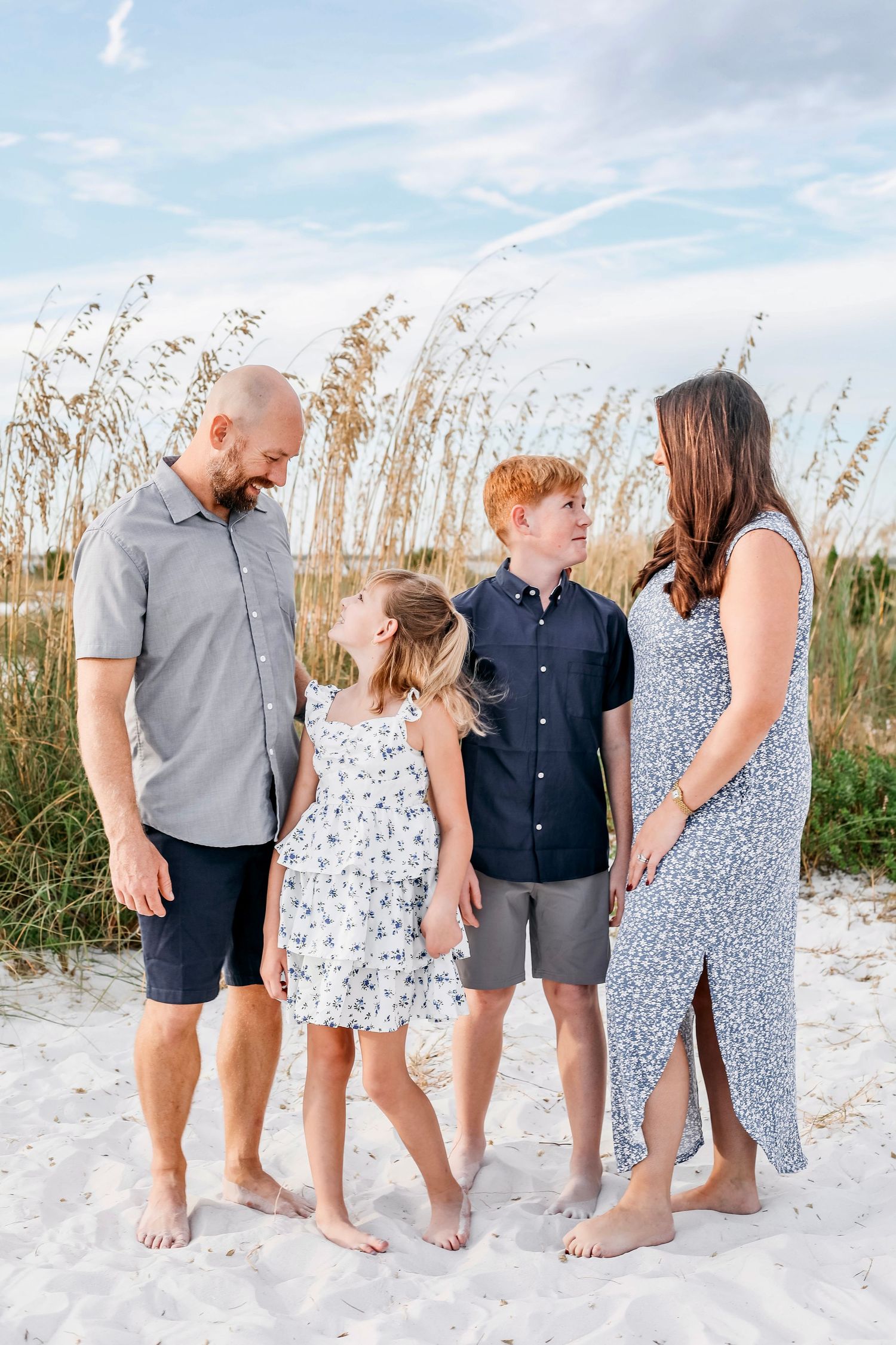 A family wearing coordinated blue and gray outfits stands together barefoot on a white sandy beach with sea oats.