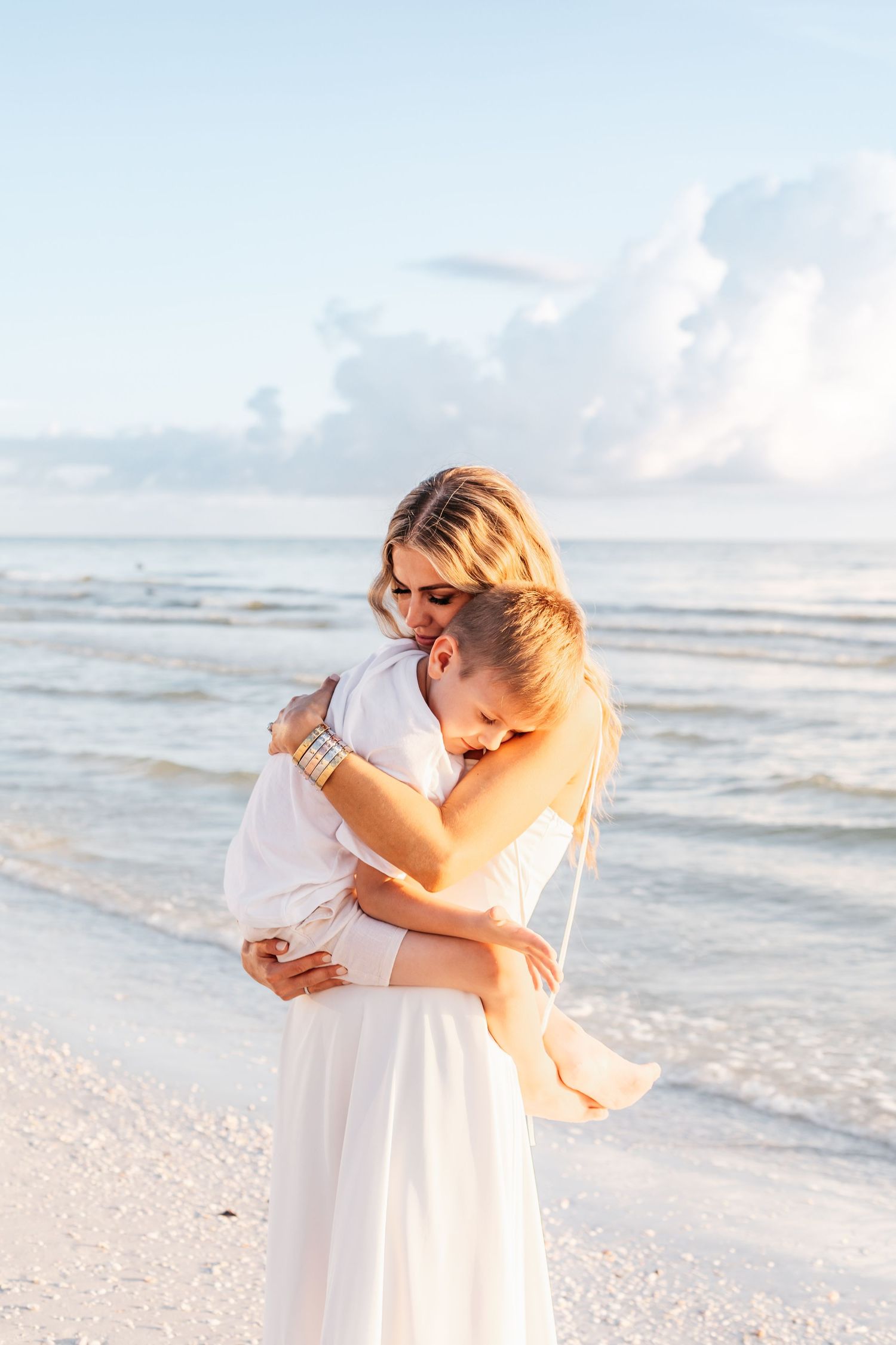 A couple embraces on a white sandy beach at sunset with gentle waves in the background.