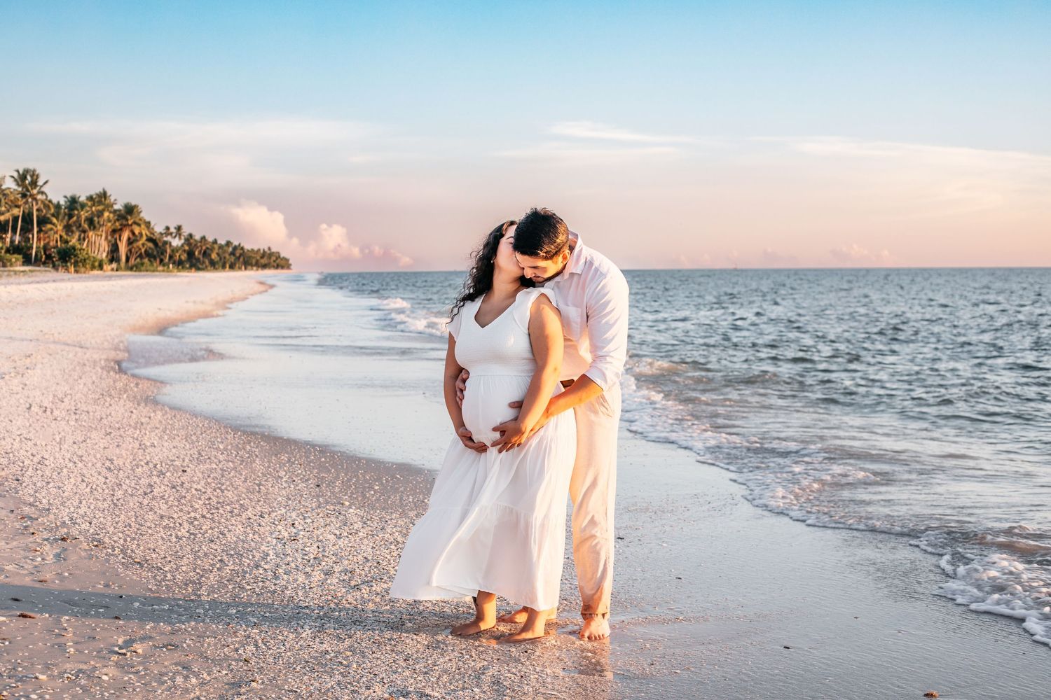 A couple in white embraces on a sunset beach with palm trees and gentle waves along the shoreline.