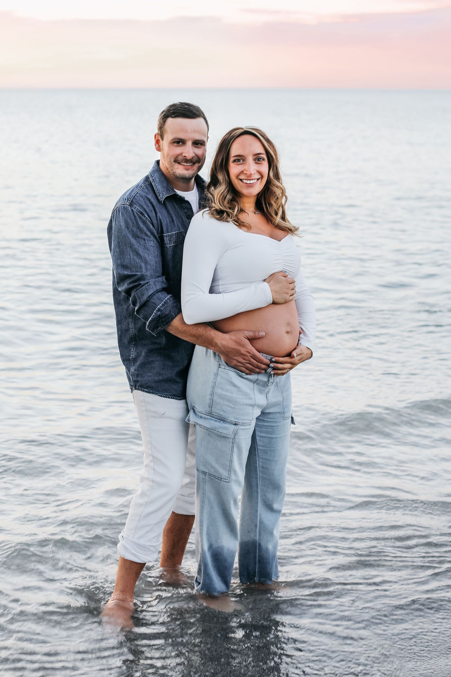 A couple stands together in shallow ocean water during sunset wearing white and blue casual clothing.