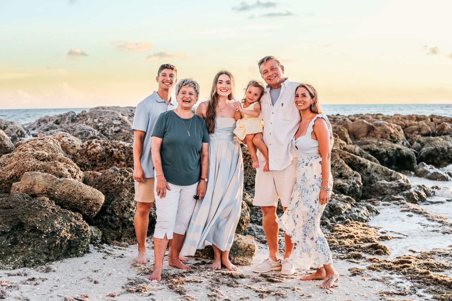 A group in coordinating beige and blue outfits poses together on a rocky beach at sunset.
